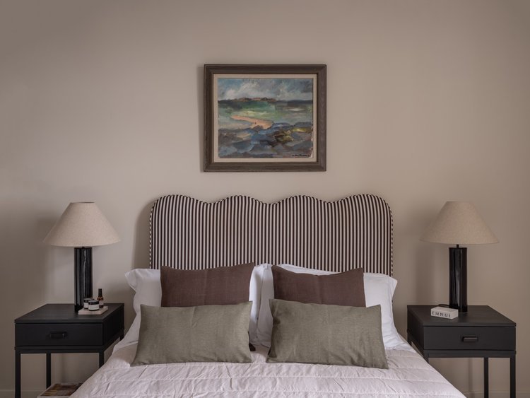 Bedroom with a striped headboard, four pillows, two bedside tables with lamps, and a landscape painting on the wall.