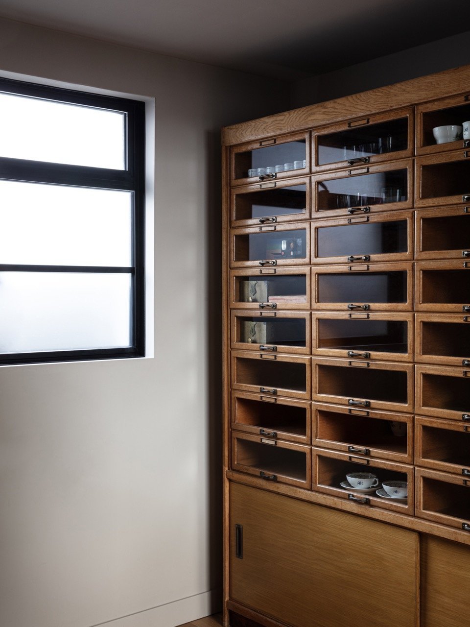 A wooden cabinet with multiple small glass-doored drawers in a room with a frosted window.