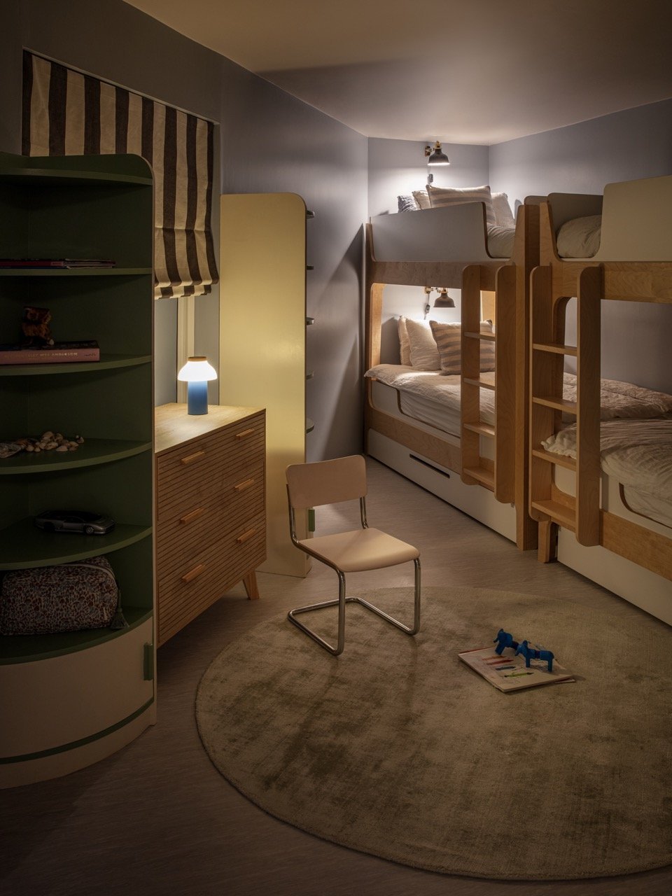 A cozy bedroom with two sets of wooden bunk beds, a beige chair, a round rug, a small bookshelf with toys and books, a wooden dresser with a lamp, and a window with a striped Roman shade.