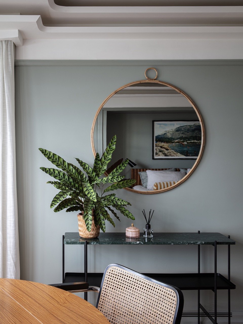 Interior of a living room with a large green potted plant on a black metal table, a round mirror on the wall, and a framed landscape painting visible in the mirror's reflection.