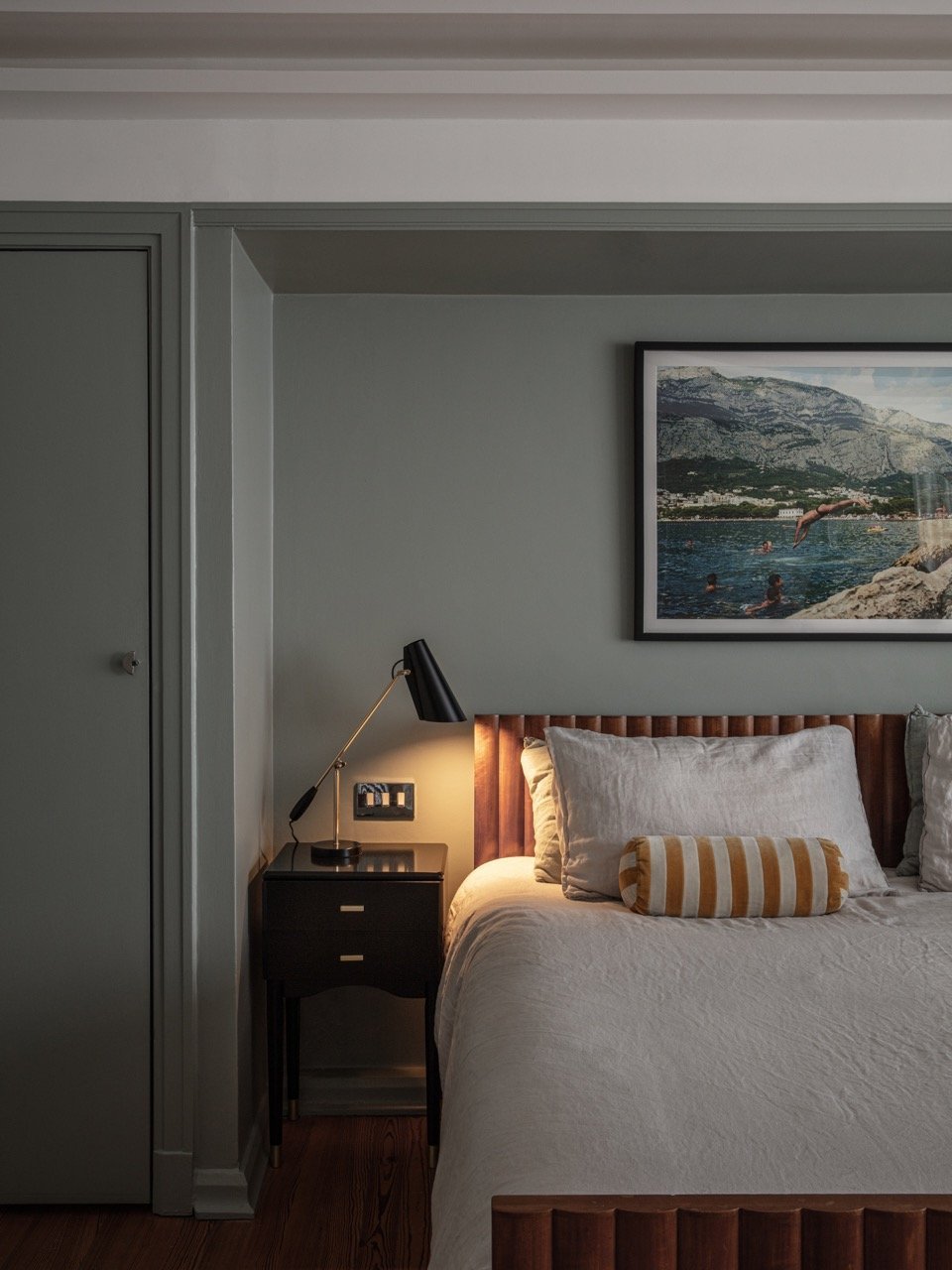 A bedroom with a bed featuring a wooden headboard, gray pillows, and a striped pillow, next to a black bedside table with a black lamp, and a framed landscape photo on the wall.