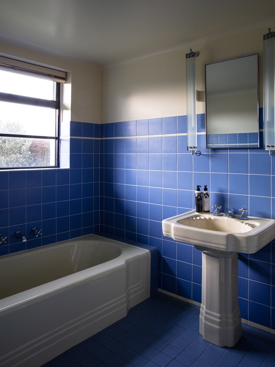 Bathroom with blue tiled walls, a bathtub, a pedestal sink with soap dispensers, a mirror, and a window showing trees outside.