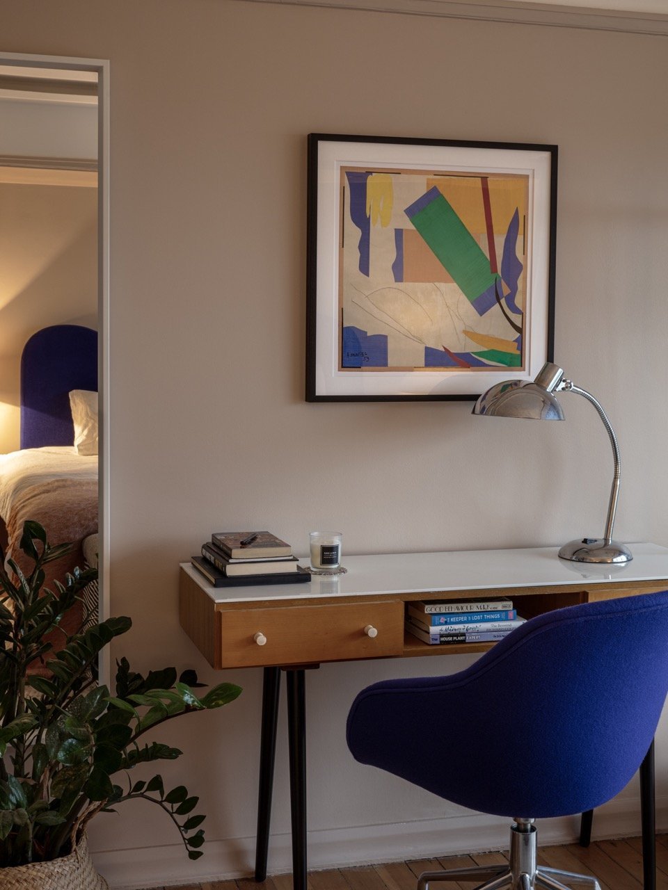 Modern workspace with a white desk, blue swivel chair, tabletop lamp, stacks of books, a candle, framed colorful abstract artwork, and a glimpse of a bedroom with a blue headboard in the background.