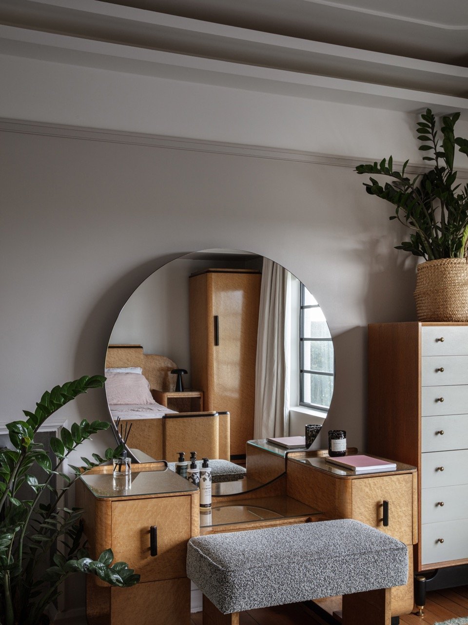 A bedroom reflected in a round mirror, showing a bed, wooden wardrobe, dresser, and window with white curtains, with plants and decor on the furniture.