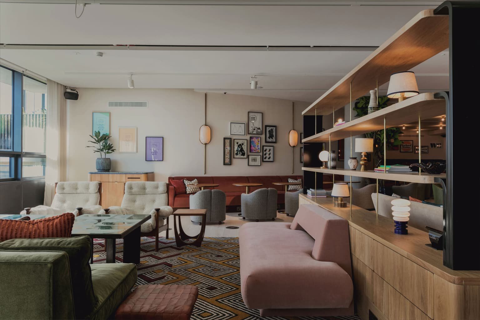 A stylish lounge with beige armchairs, a pink sofa, and a pattern rug. A wooden partition with shelves holds lamps and decorative items. The back wall has framed artwork and wall-mounted lights, with large windows allowing natural light in.