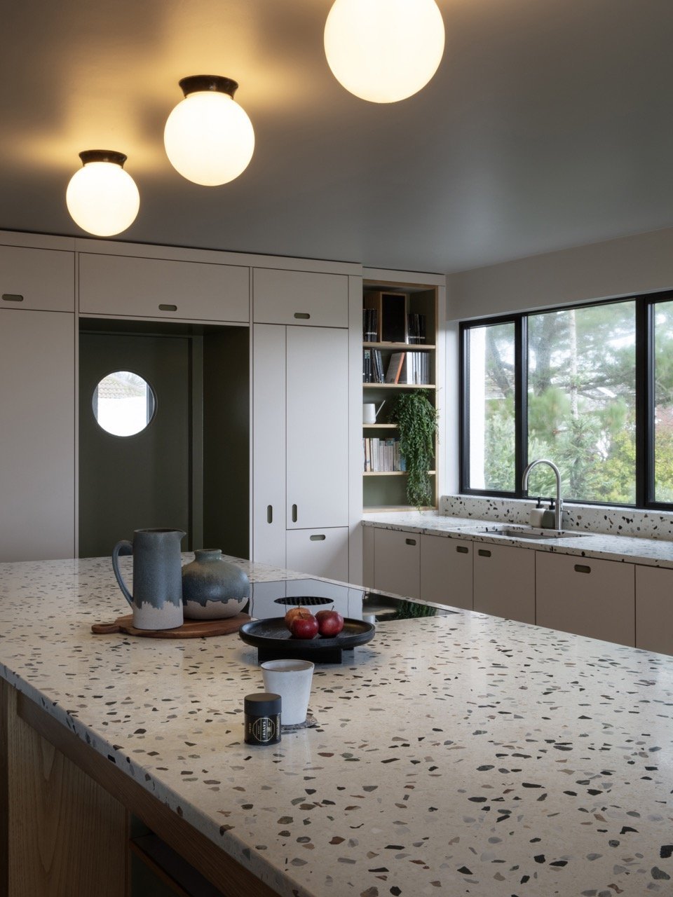 Modern kitchen with terrazzo countertop, white cabinets, window overlooking trees, and three ceiling lights.