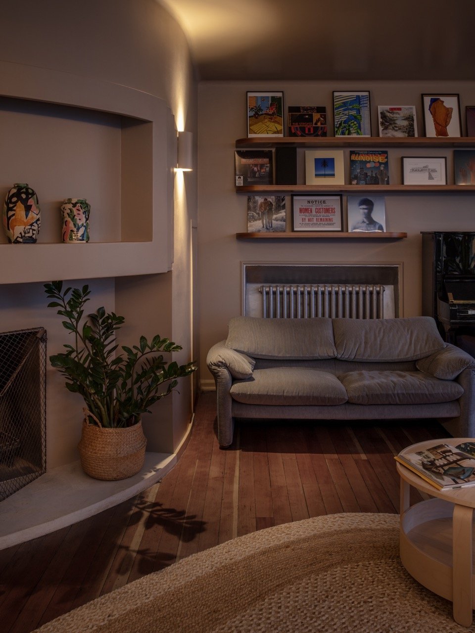 Living room with a gray sofa, wooden floors, two wooden shelves with framed art and photographs, potted plant in a woven basket, and a small round coffee table