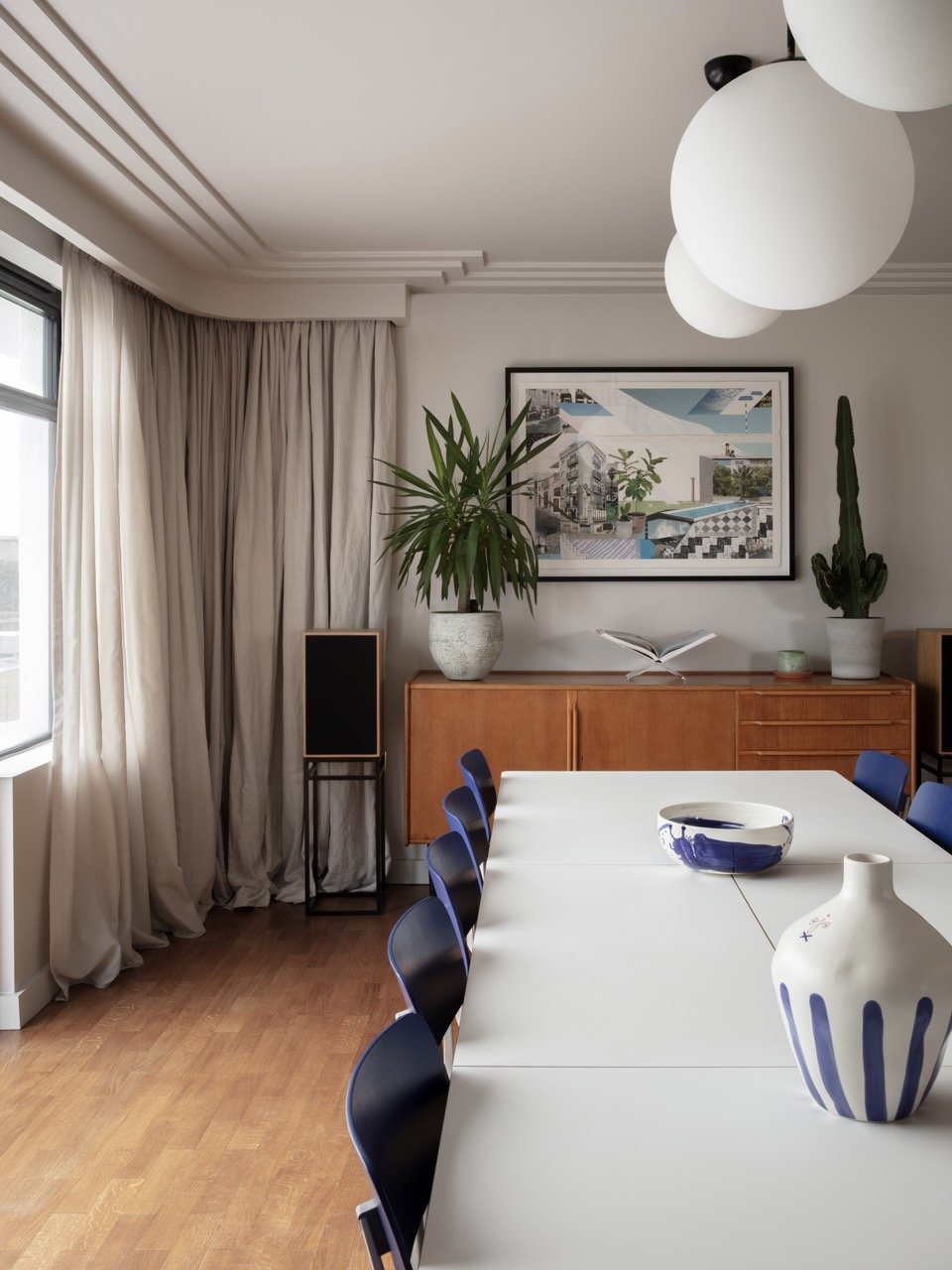 Modern dining room with white table, blue chairs, potted plants, framed artwork, large window with beige curtains, and hanging white spherical light fixtures.