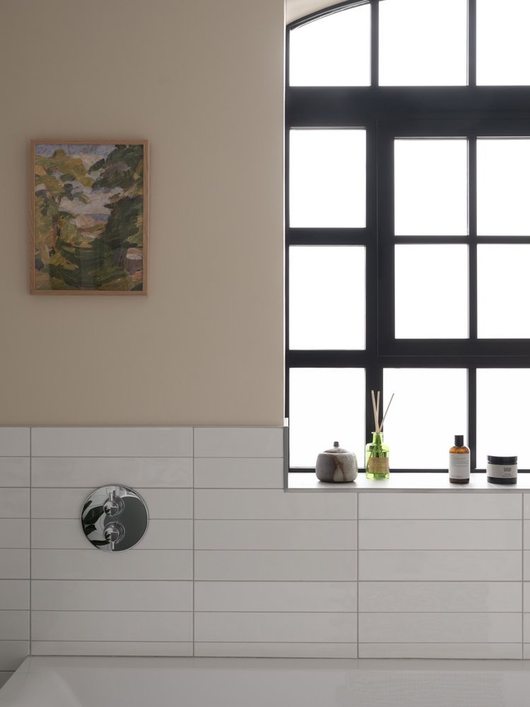 A bathroom with white tiled walls, a black-framed window, and a windowsill holding a stone, green reed diffuser, and bath products. A framed painting hangs on the beige upper wall.