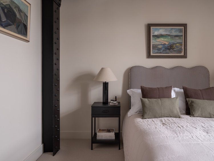 A bedroom with a bed that has striped headboard, decorative pillows, a black nightstand with a lamp and books, and two framed landscape paintings on the walls.