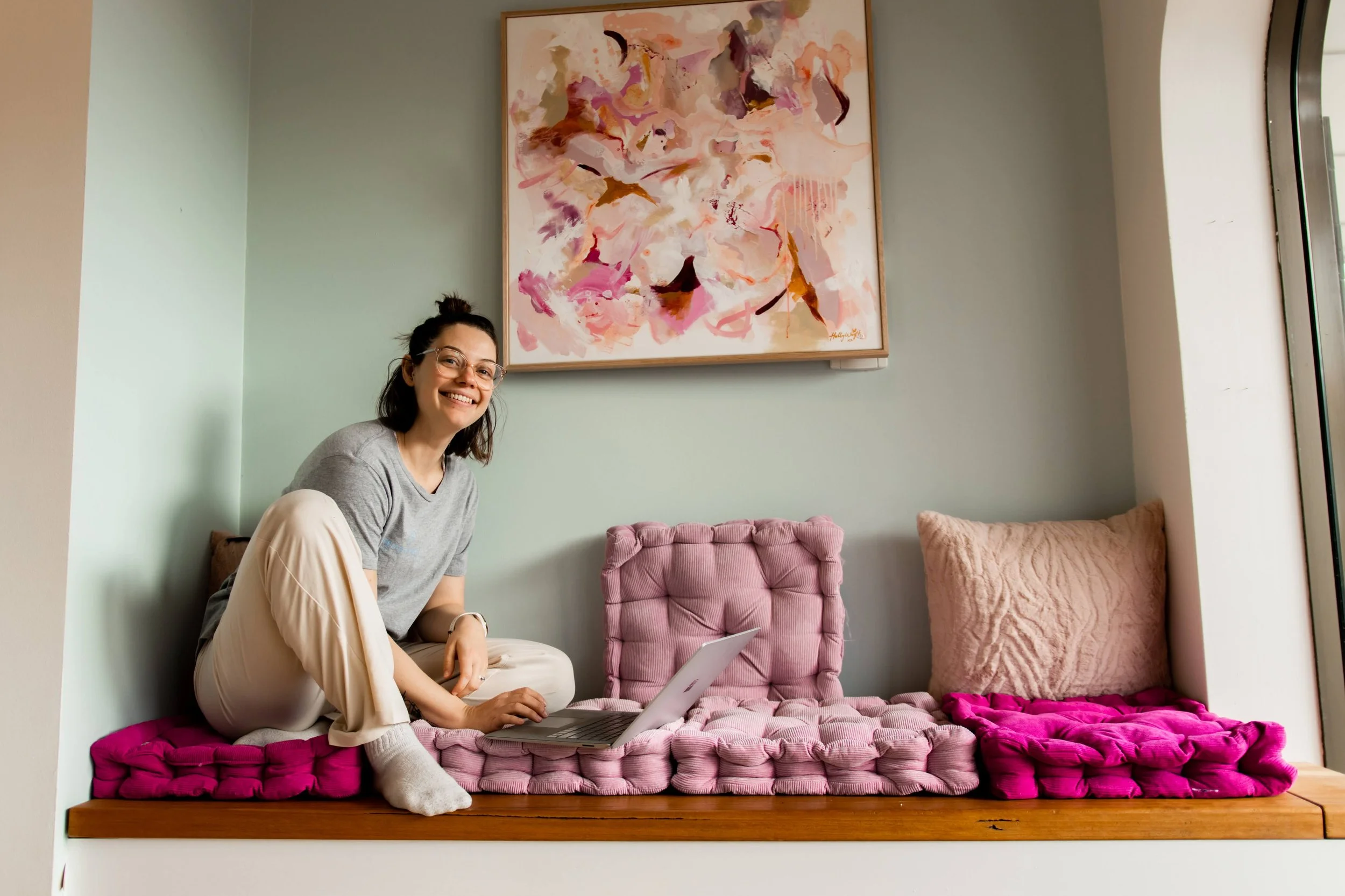 A woman with glasses sitting on a pink velvet window seat, using a laptop, smiling at the camera, with abstract artwork hanging above her and cushions around her.
