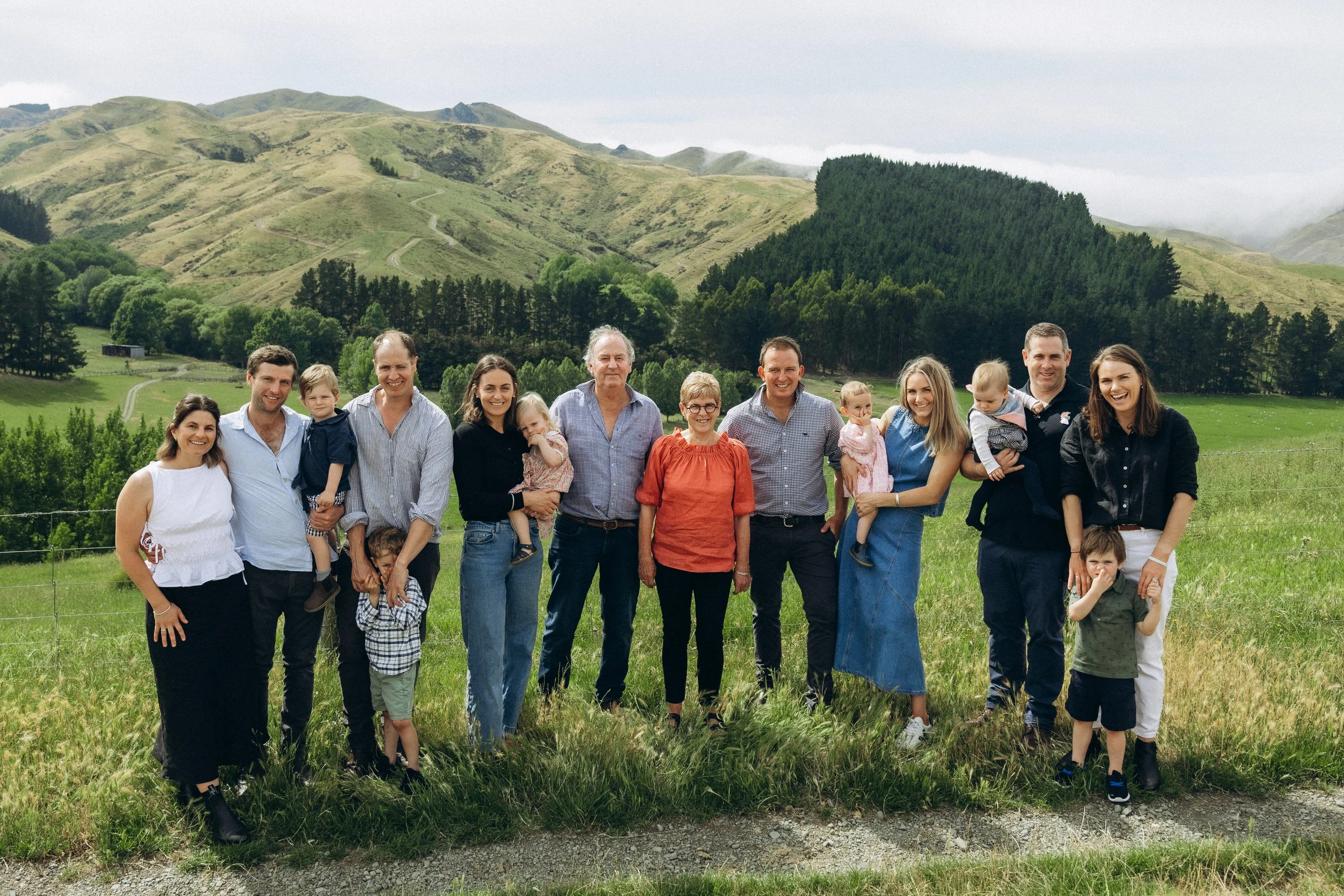 A group of people posing outside in front of a hilly landscape. The group includes men, women, and several children. They are standing on grass with mountains and trees in the background.