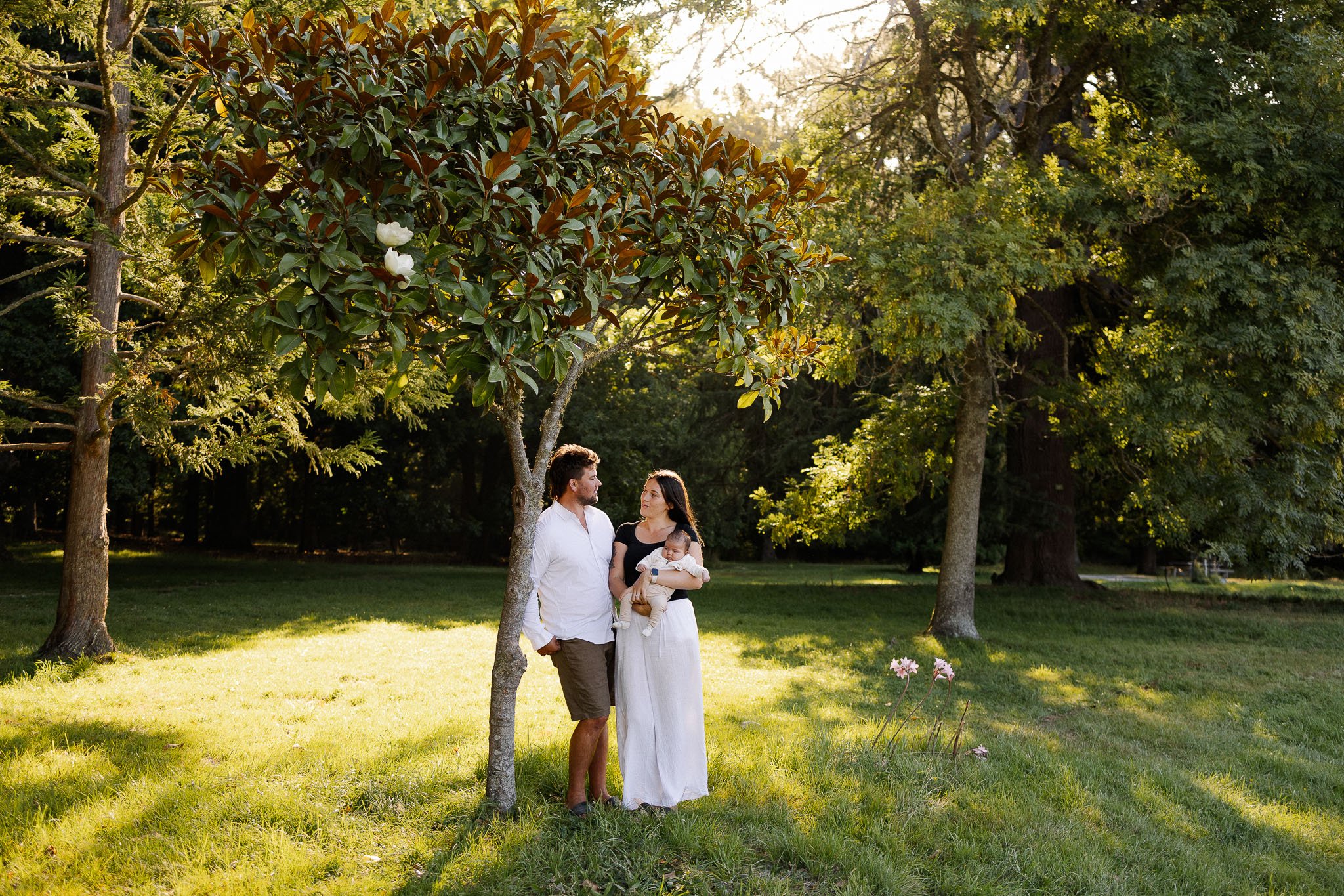 A family of three standing under a large tree in a park during daytime. The man is wearing a white shirt and shorts, the woman is wearing a white dress and holding a baby, and they are looking at each other. The background features green grass, other trees, and sunlight filtering through the leaves.