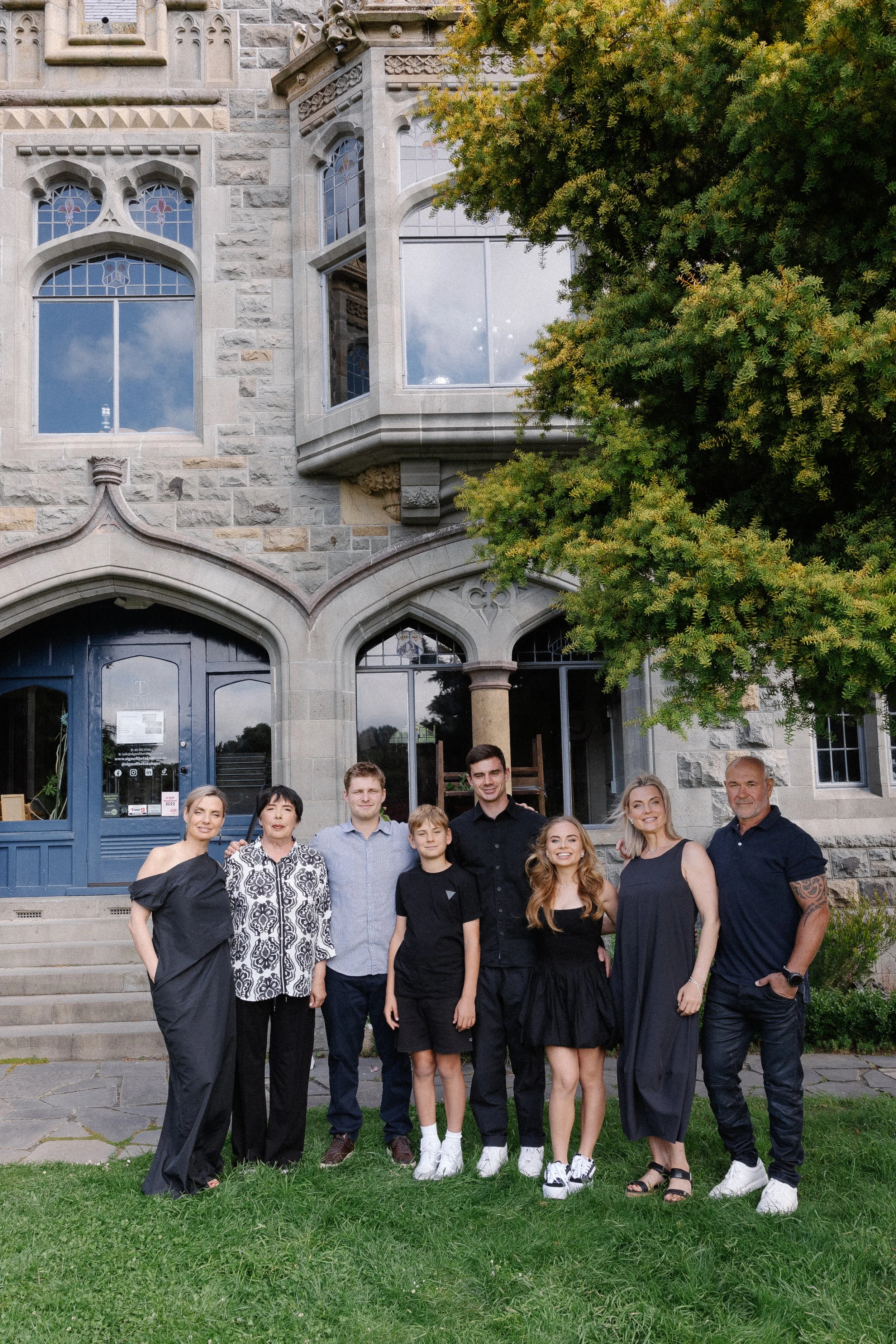A group of people posing outdoors in front of a historic building with large windows and stone architecture. They are standing on a lawn, with a tree to the right. The people are dressed in semi-formal attire.
