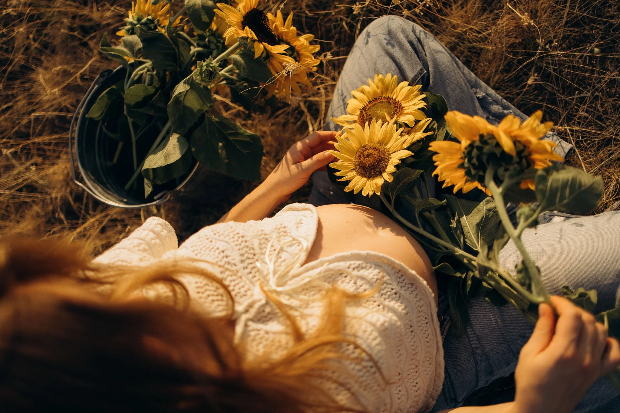 Person sitting on the ground in grass, holding yellow sunflowers while wearing jeans and a cream-colored knit sweater.