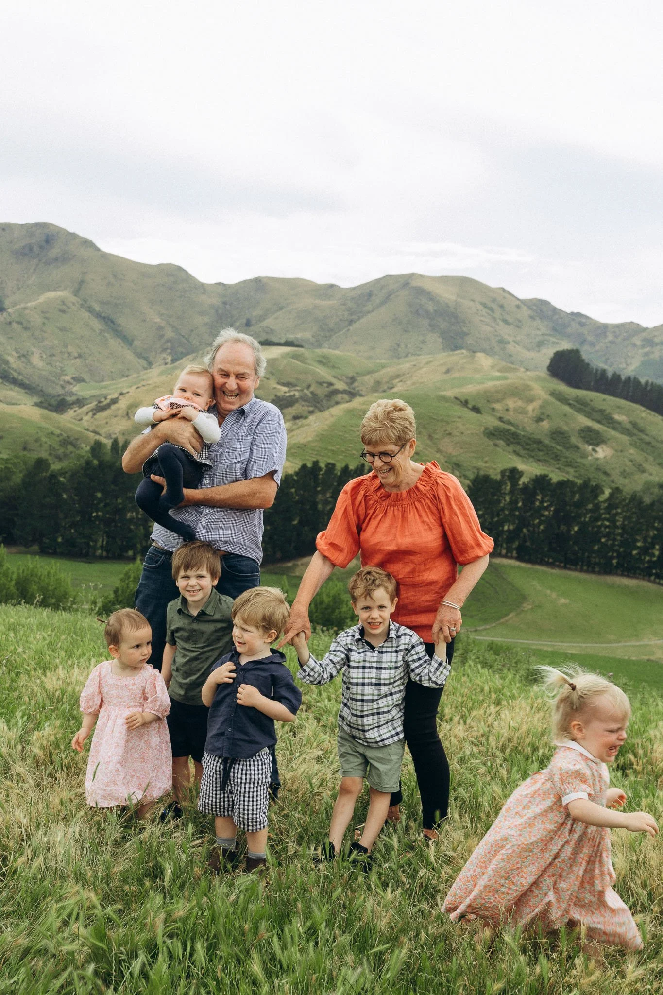 A multi-generational family enjoying a day outdoors in a lush green field with rolling hills in the background. The scene includes three adults and six children, some holding hands, smiling and playing.