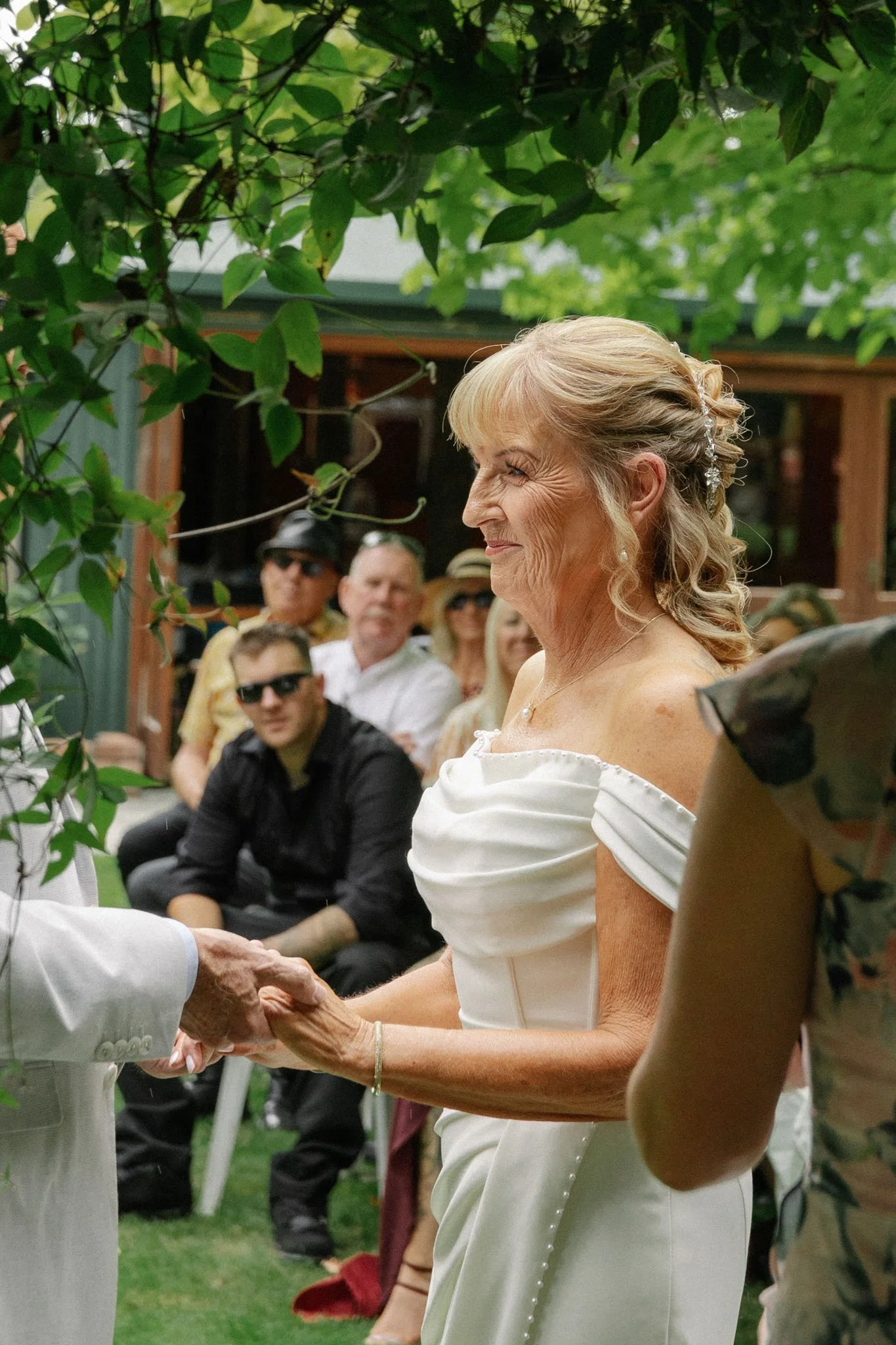 An elderly woman in a white dress during a wedding ceremony, holding hands with a person dressed in white, with guests seated in the background.