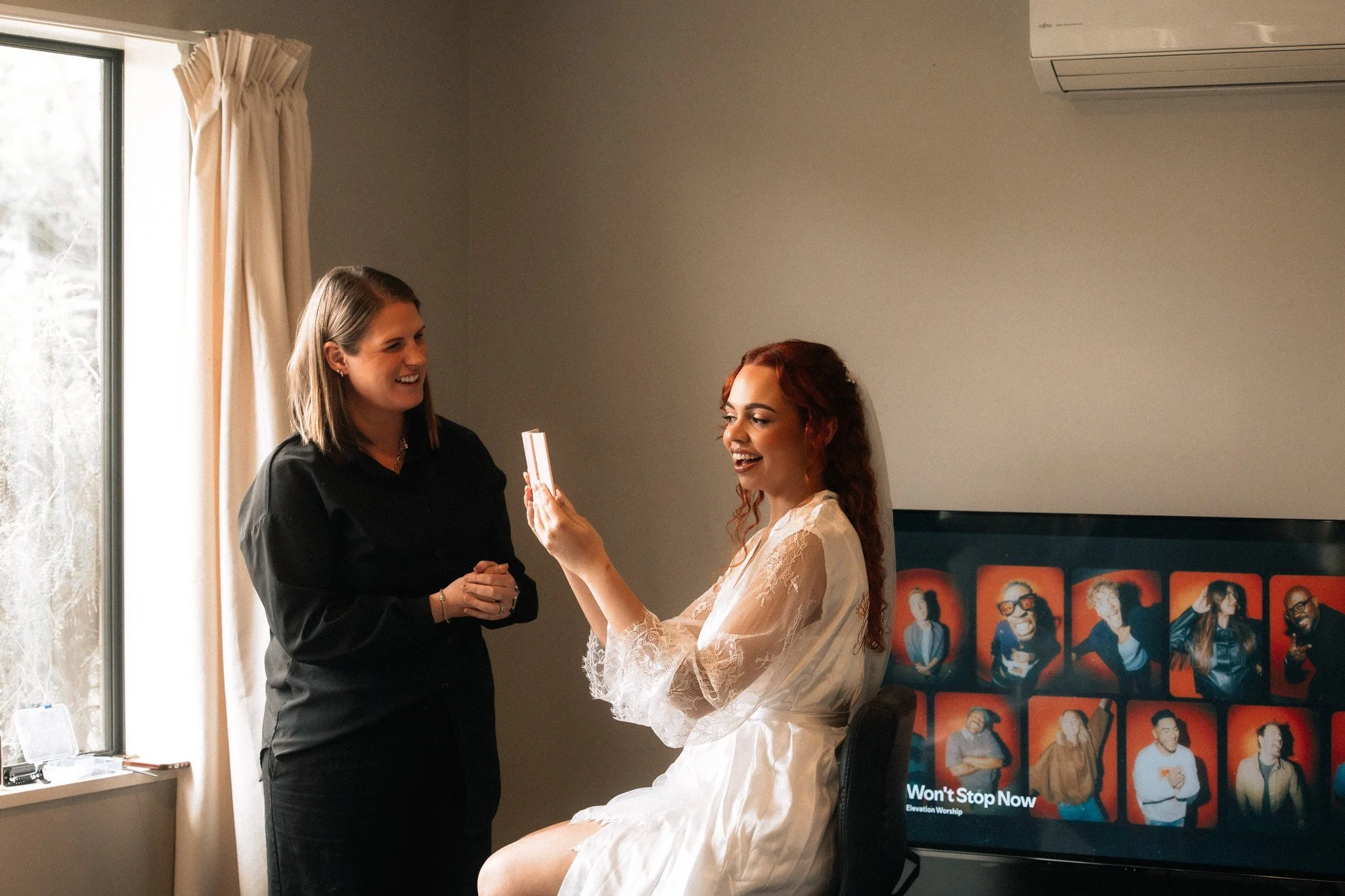 Two women in a room, one sitting on a chair and the other standing, exchanging a small rectangular object, possibly a gift or card, near a large window with cream curtains, with a flatscreen TV or digital display in the background showing portraits of various people.