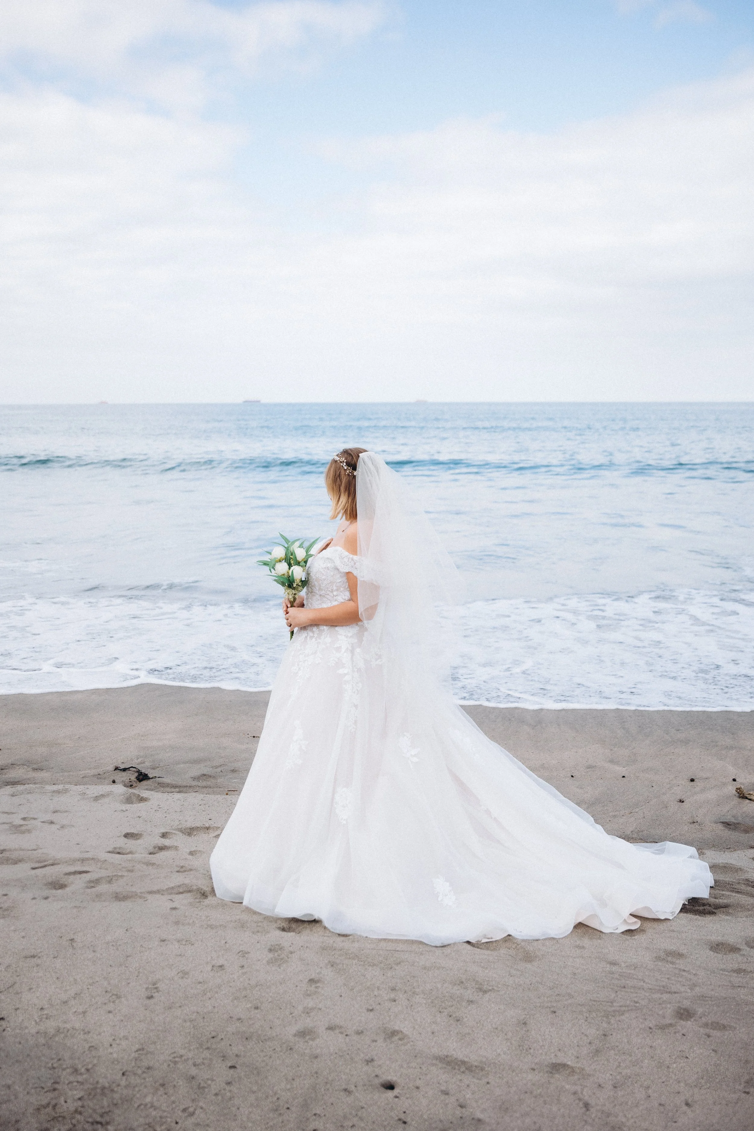 Bride in wedding dress holding flowers, standing on sandy beach with ocean in background.