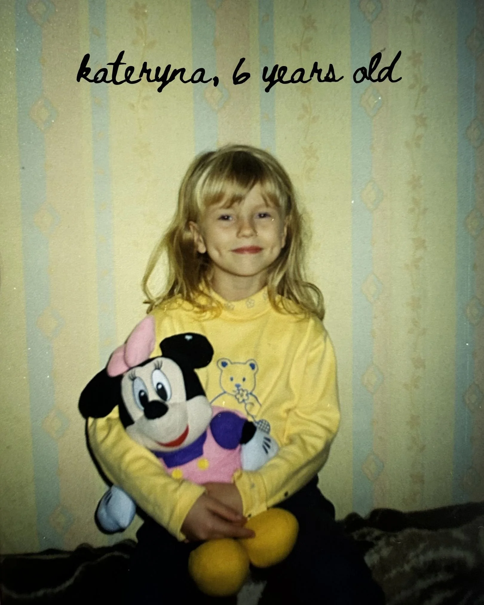 A young girl with light brown hair, wearing a yellow long-sleeve shirt with a bear and a lion design, is sitting against a light-colored wall with a subtle patterned wallpaper. She is smiling slightly and holding a plush Mickey Mouse toy. Two tennis balls are on her lap.