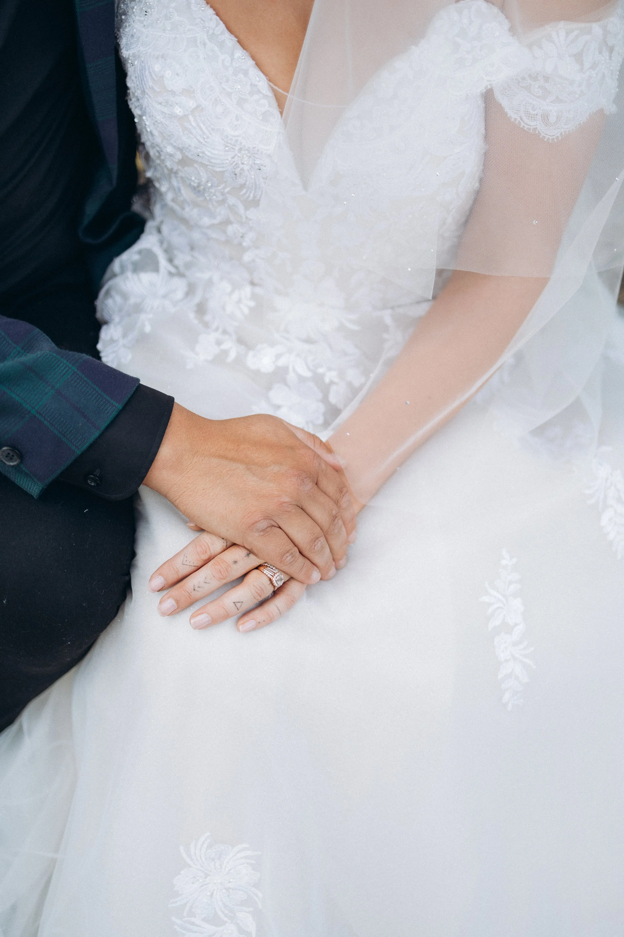 Close-up of a married couple's hands on a lace wedding dress, with geometric tattoos and a wedding ring visible.