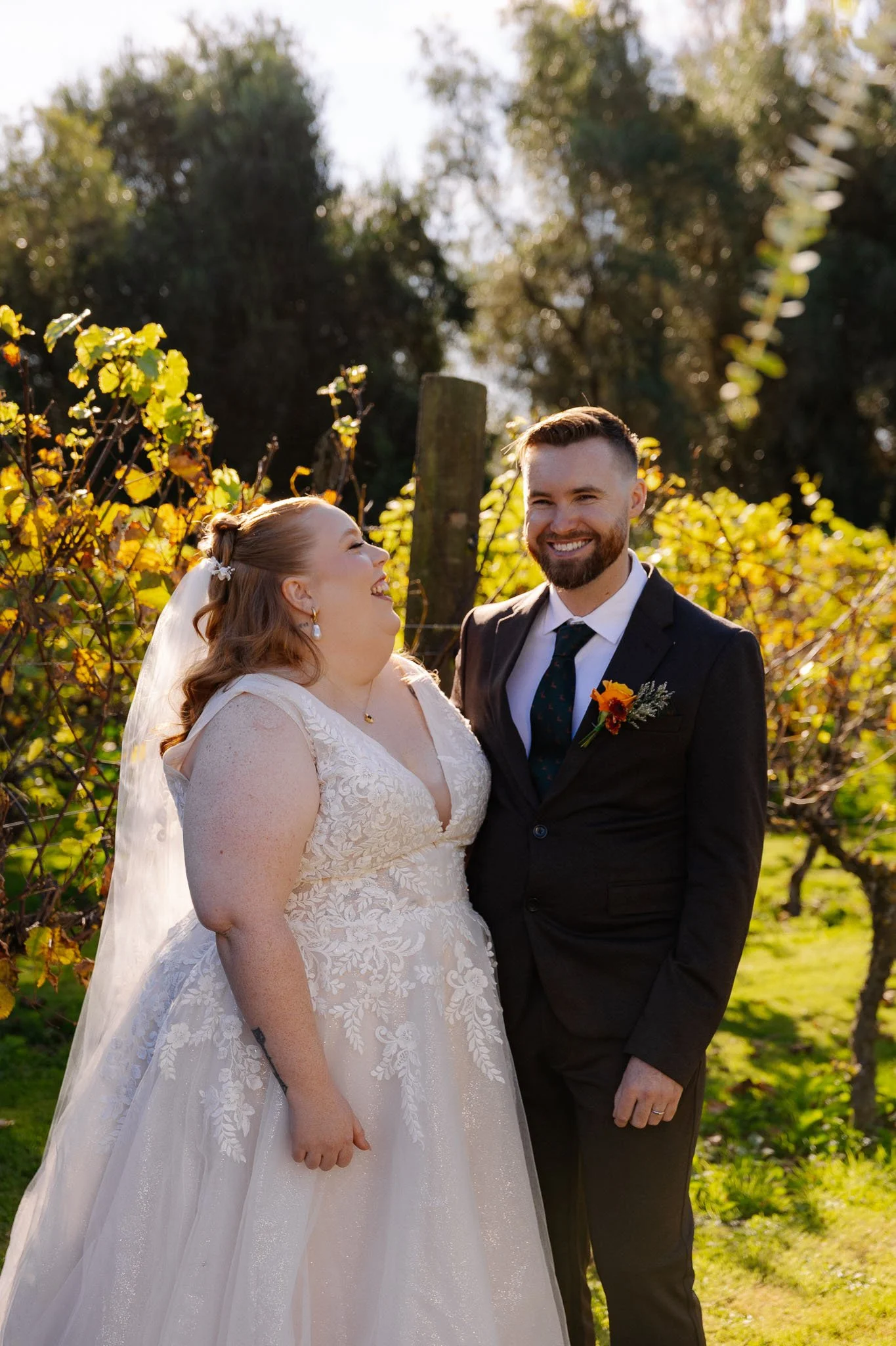 A bride and groom smiling and looking at each other outdoors with trees and greenery in the background, during a wedding.