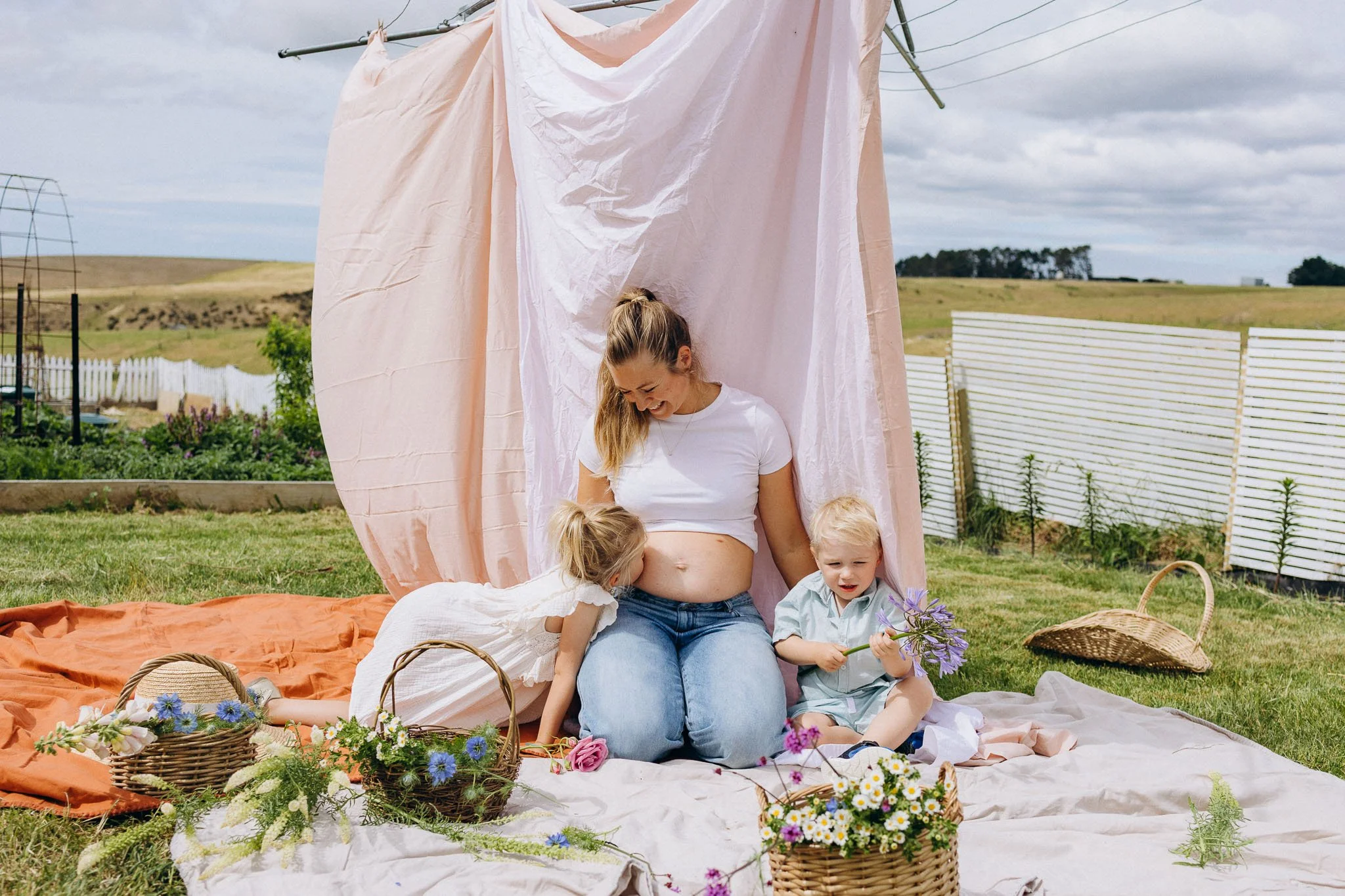 A pregnant woman sitting on a blanket outdoors with three young children, flowers, baskets, and a pink fabric backdrop, in a field with a white fence and cloudy sky.