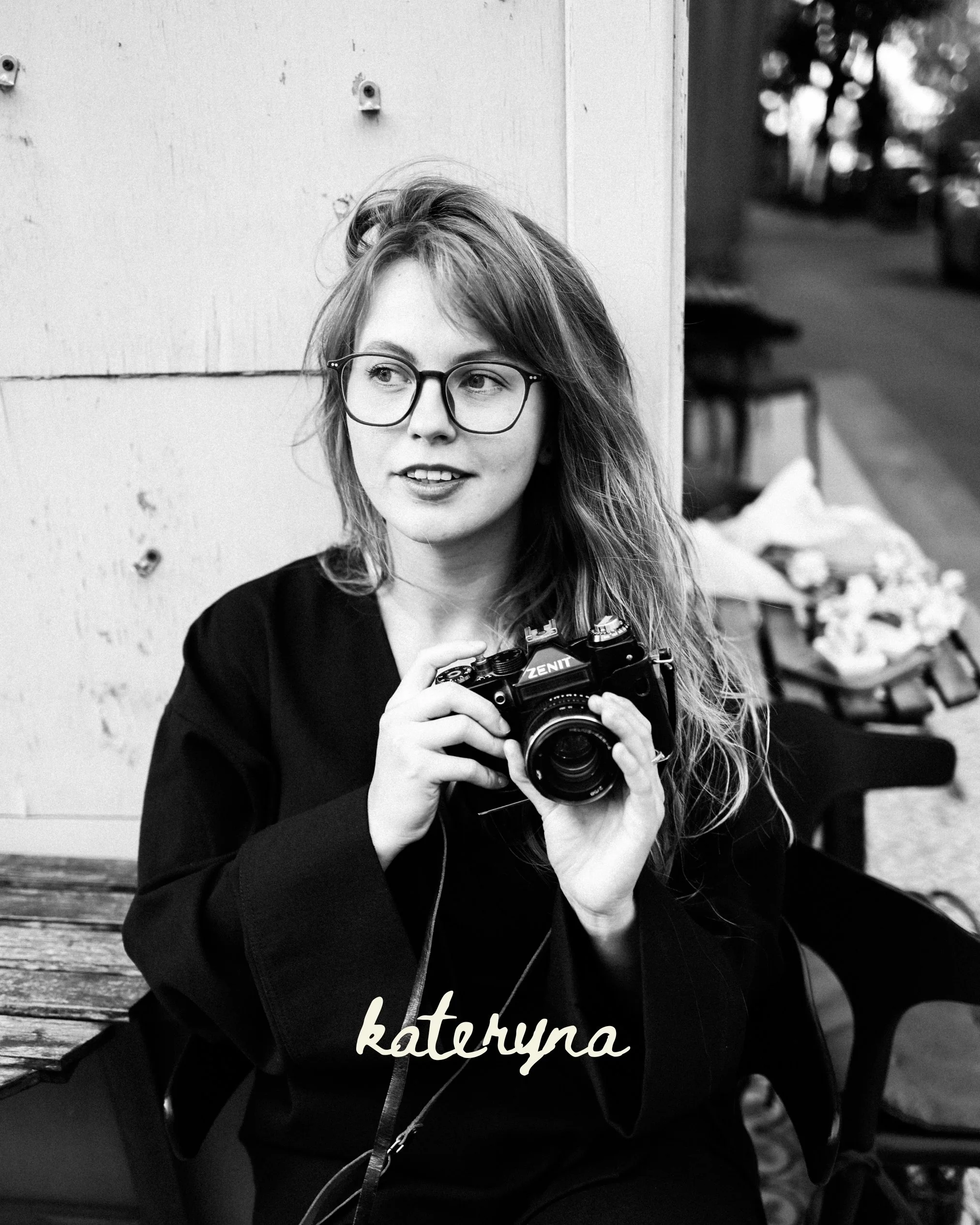 A young woman with glasses holding a camera, sitting on a bench, with a blurred background of trees and a street, and text in Polish that says 'fotografia kateyma.'