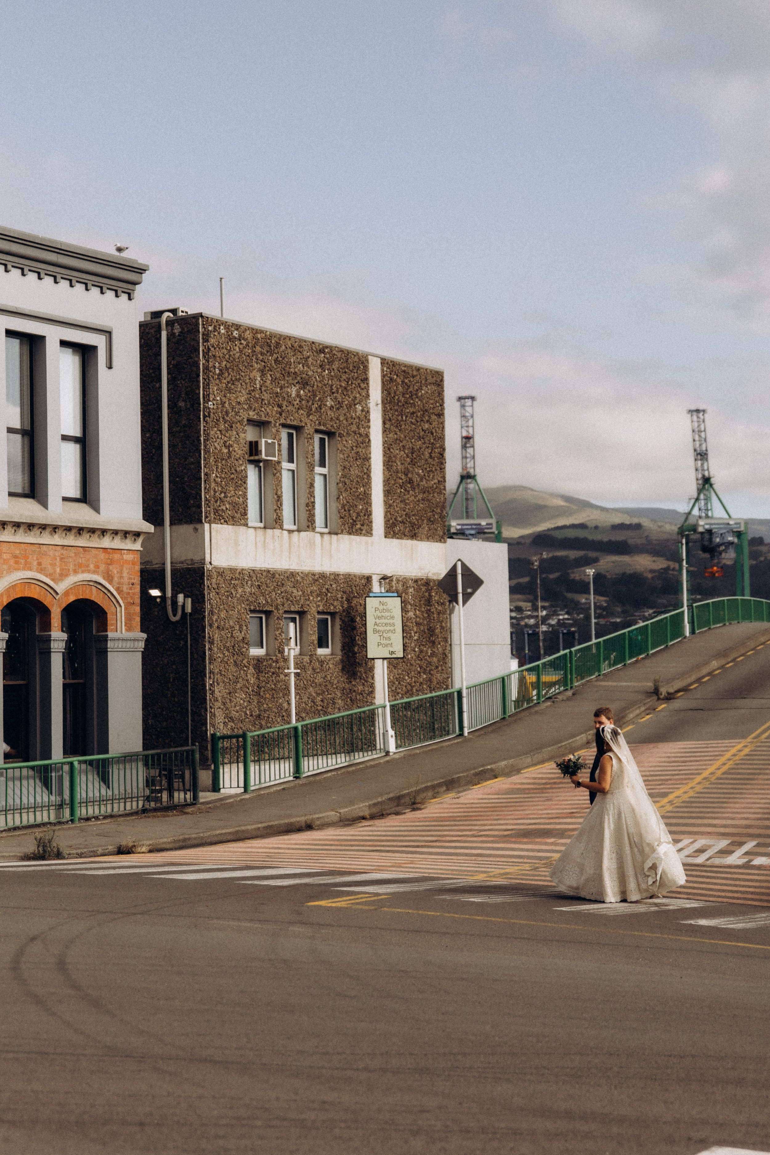 Bride in wedding dress crossing a street near urban buildings and cranes in the background.