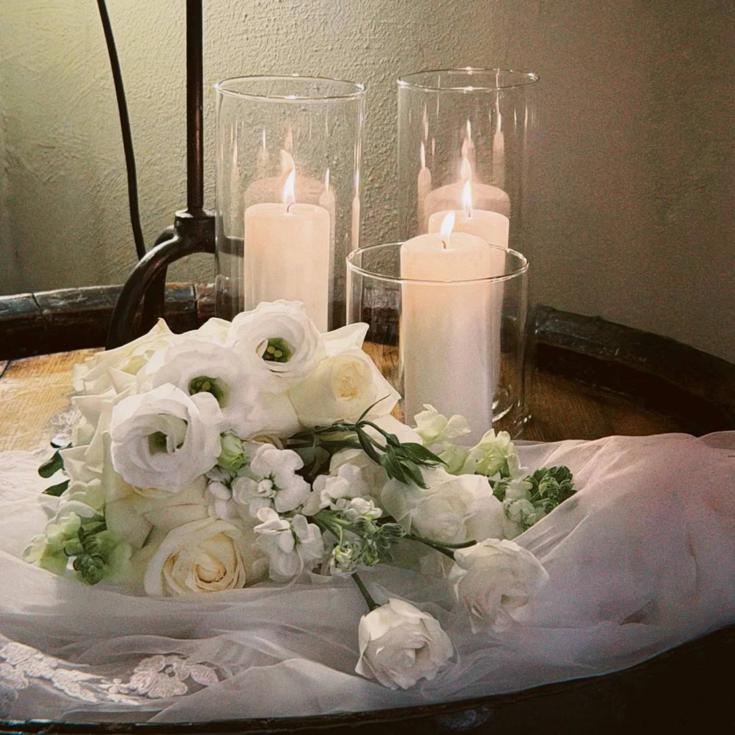 A bouquet of white flowers with white roses and greenery, placed on a table. Behind it are three lit white candles in glass holders, with additional glass holders in the background. The setting appears to be decorative and possibly for a special occasion.