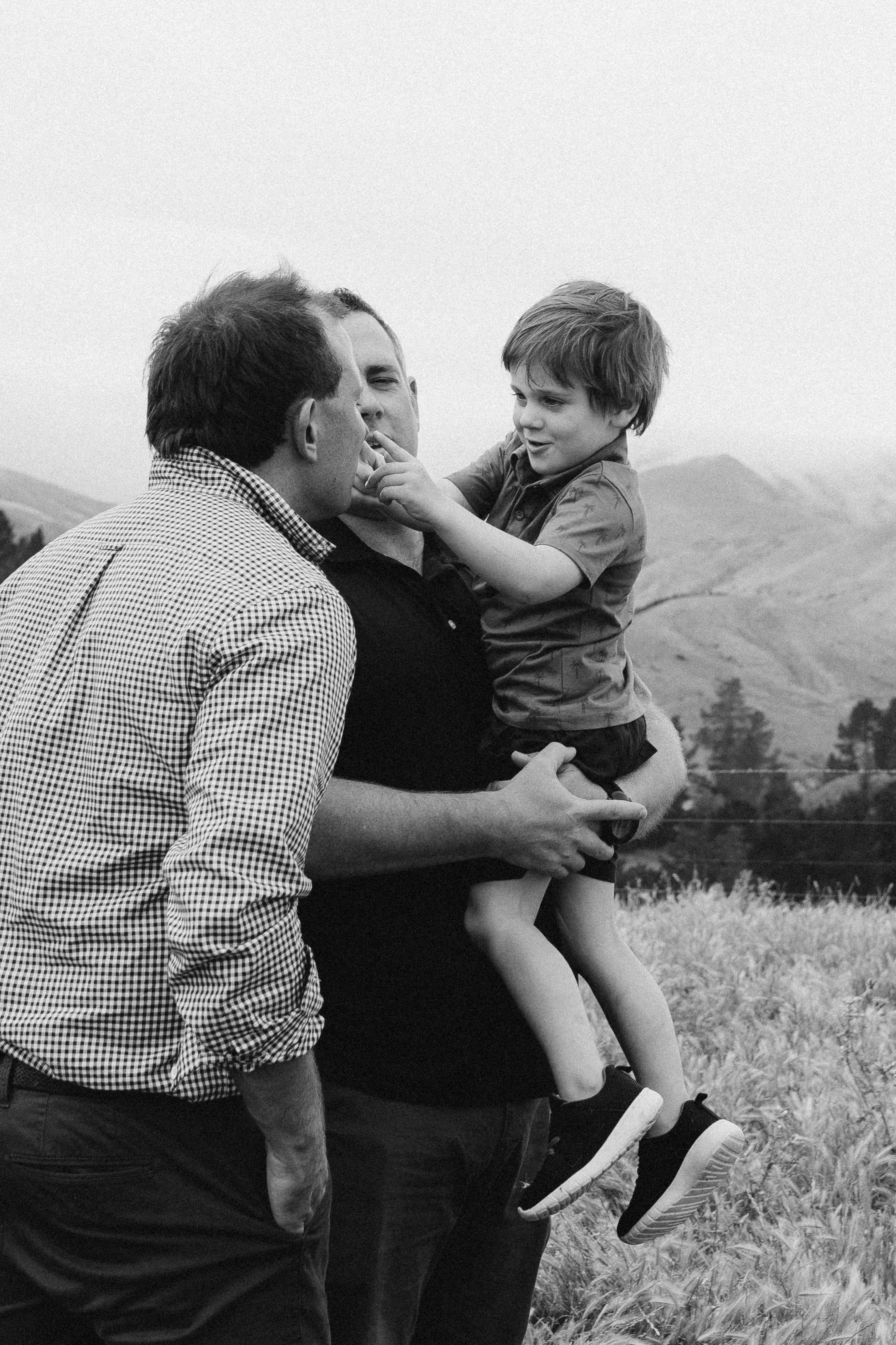 A black and white photo of two men with a young boy in an outdoor setting. One man is holding the child, who is playfully touching the other man's face. The landscape includes hills and trees in the background.