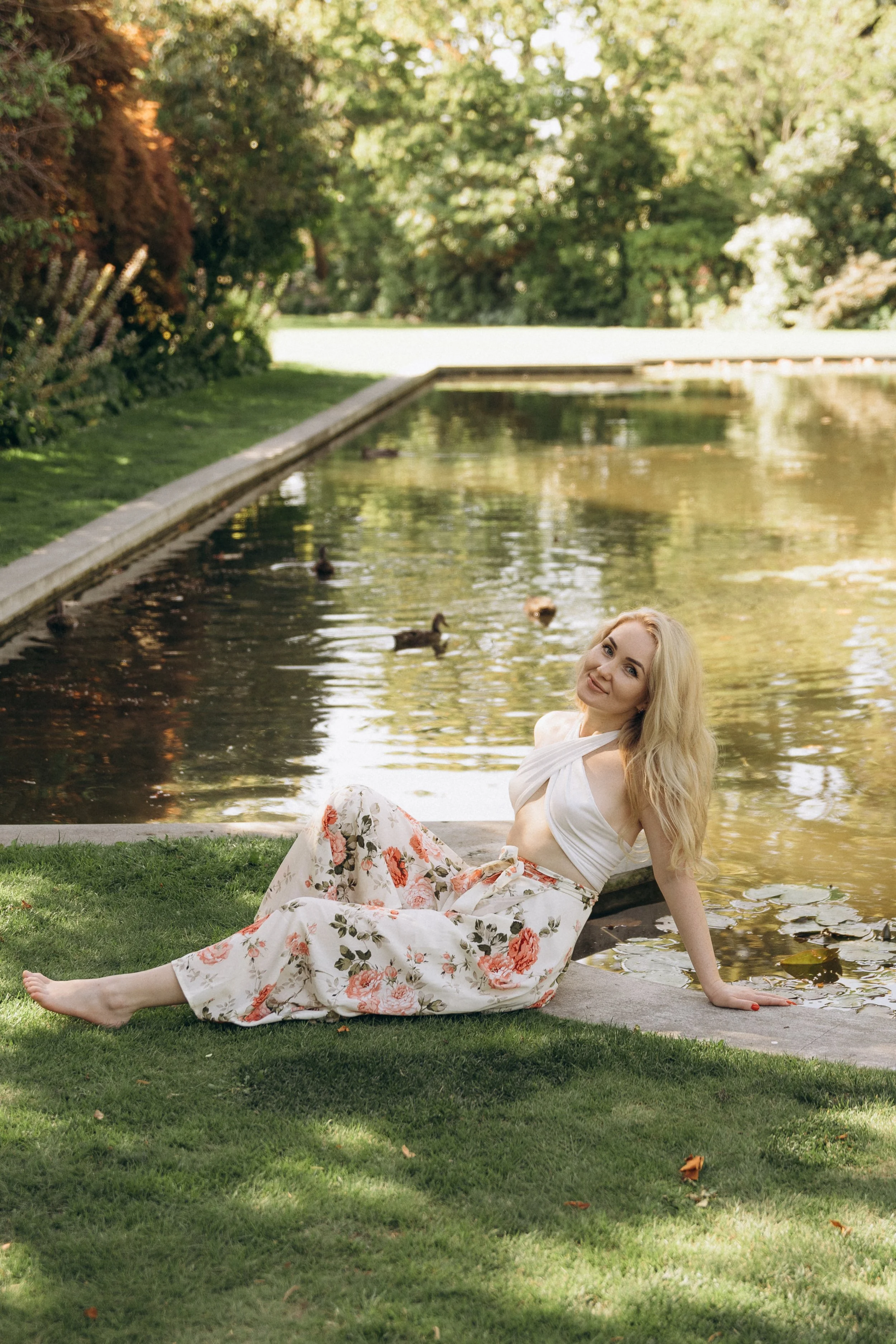 A woman with long blonde hair, wearing a white top and floral skirt, sitting on grass near a pond in a garden.