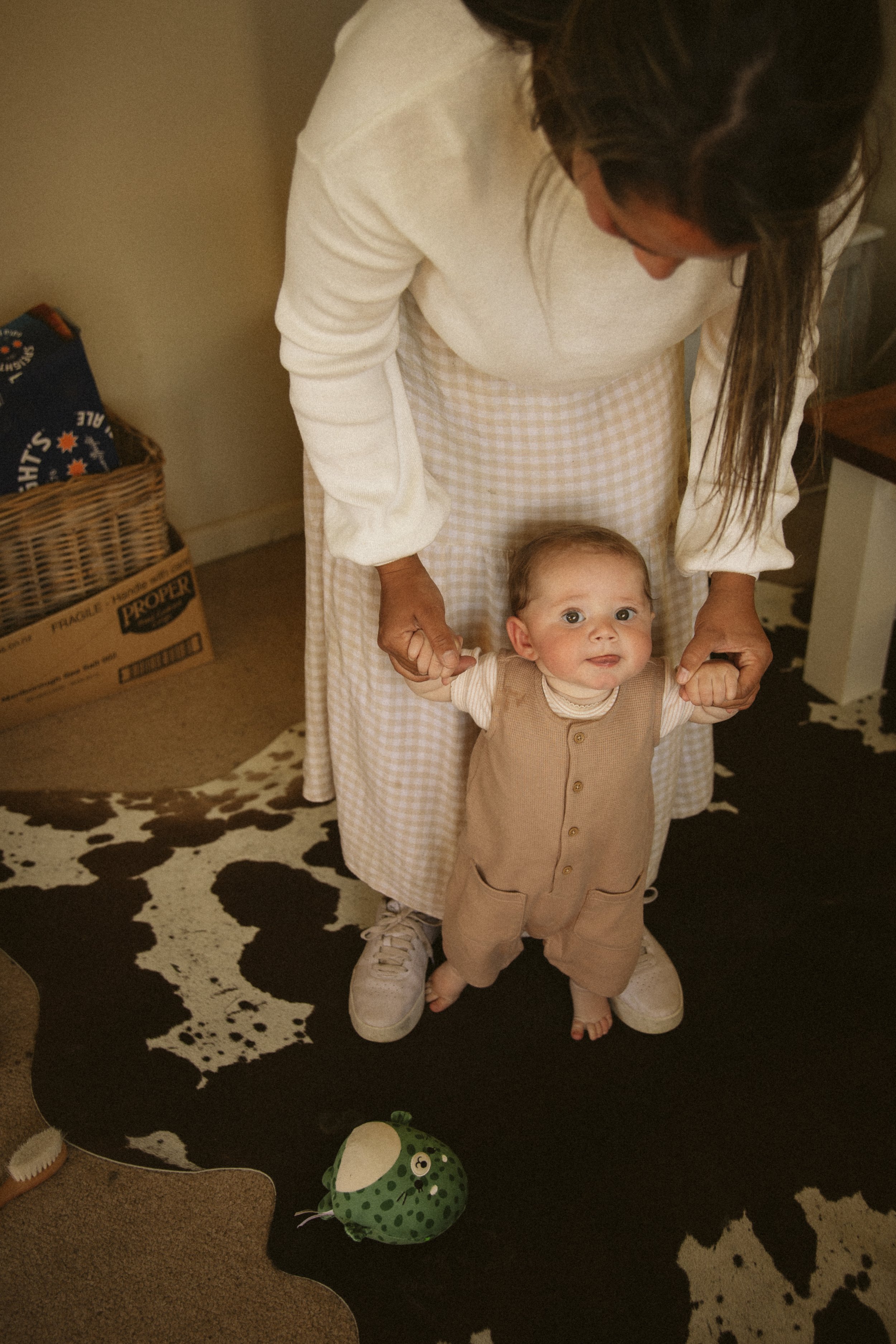 A baby wearing a beige outfit, supported by an adult holding the baby's hands, standing on a cowhide rug. A green toy is on the floor nearby.