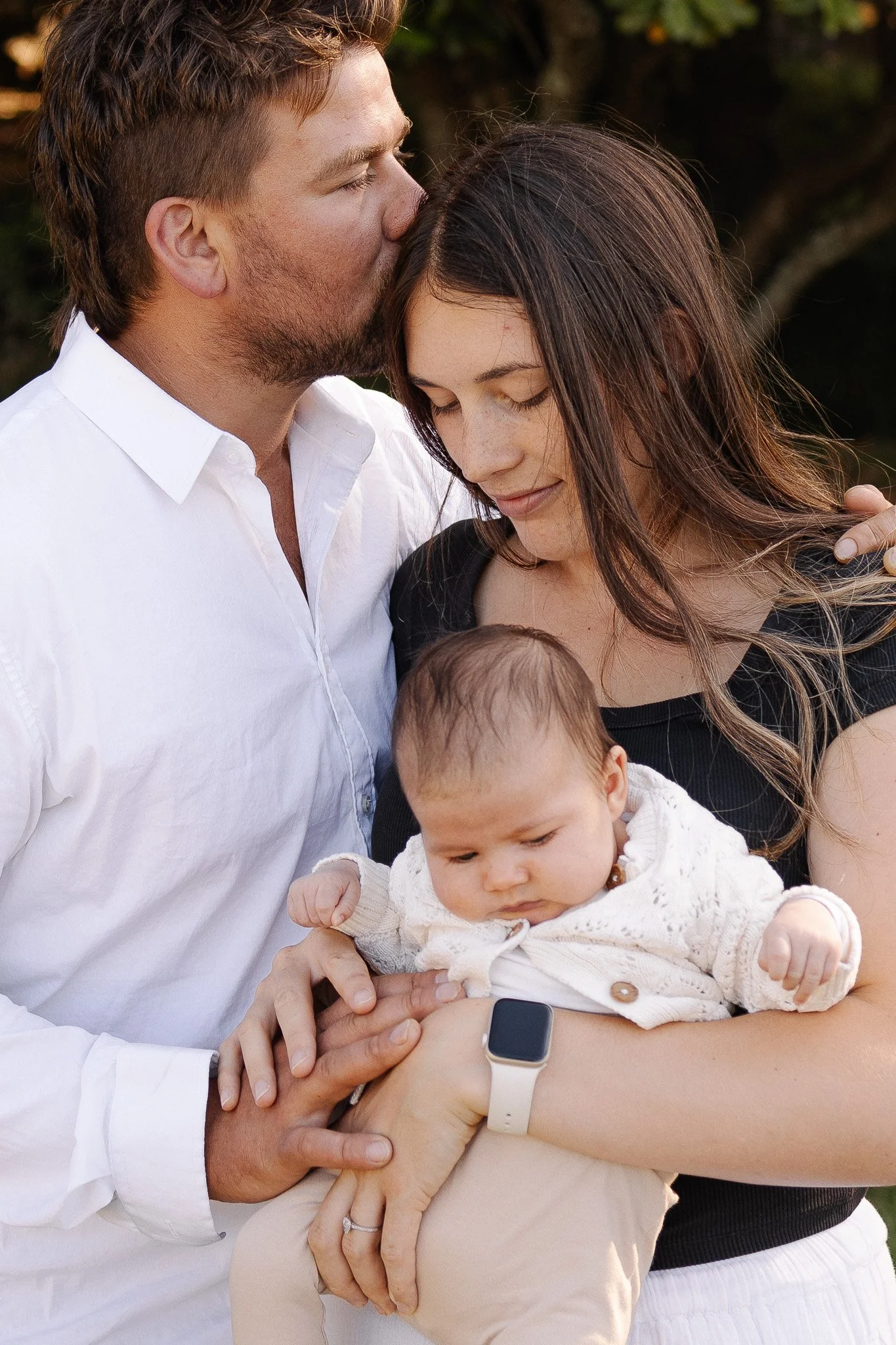 A loving couple holds their baby, with the man kissing the woman's forehead. The baby is wearing a white sweater. The woman has an Apple Watch on her wrist.