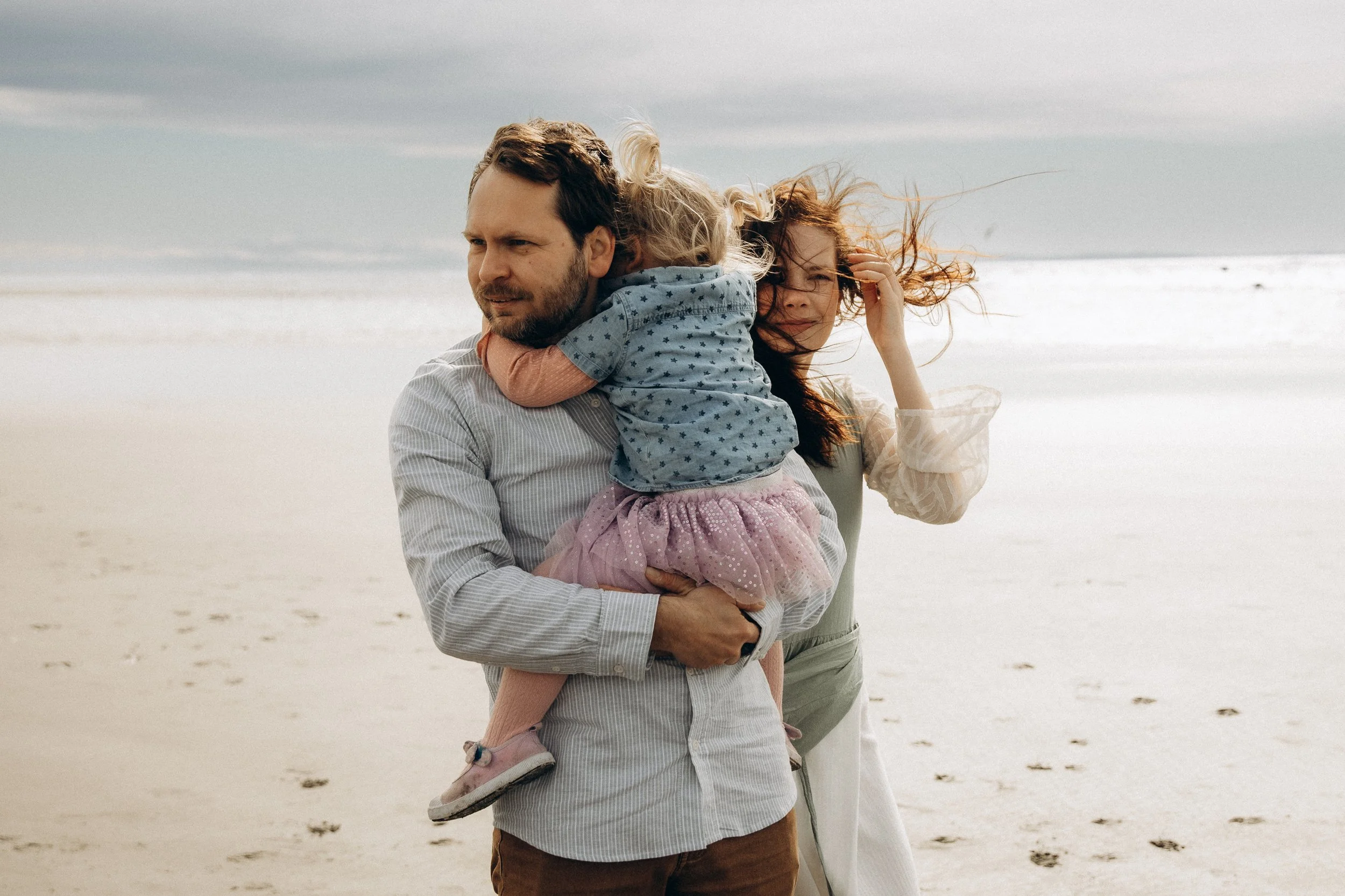 Family on a windy beach, man holding a child in a pink skirt, woman adjusting hair.