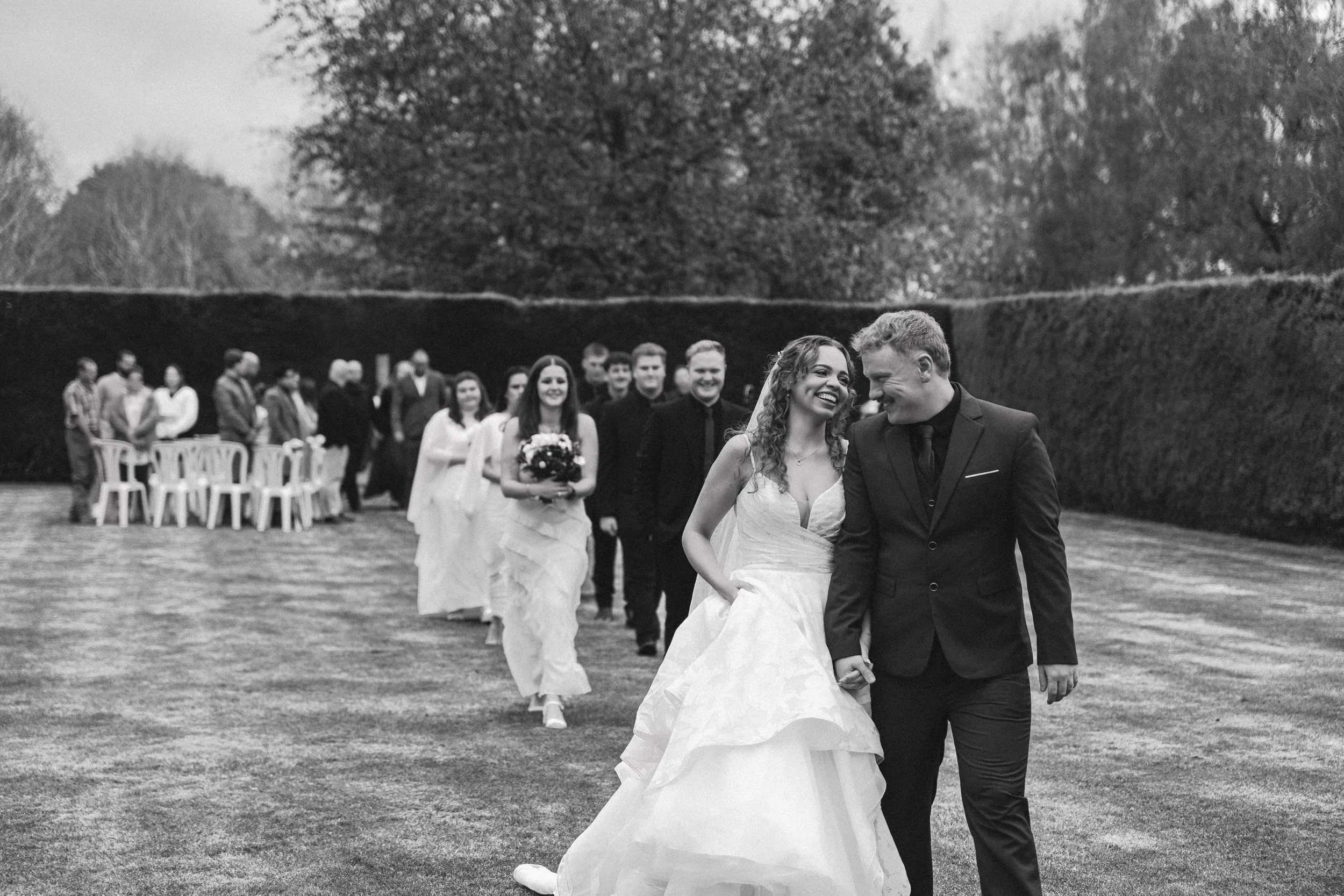 A wedding couple walking hand in hand outdoors, with the bride in a white gown and the groom in a black suit, smiling and looking at each other, surrounded by wedding guests standing and sitting in the background.