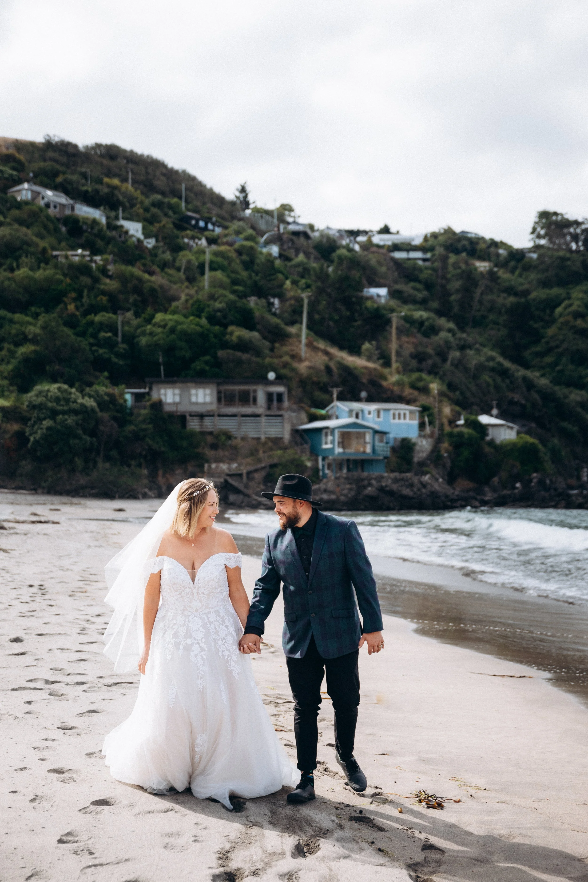 Bride and groom walking on beach, hillside houses in background