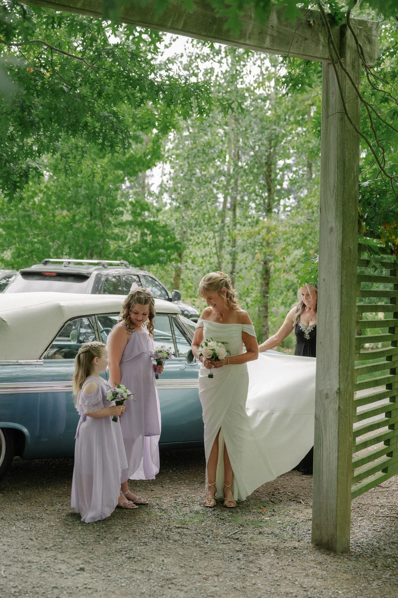 A wedding scene outdoors with a bride in a white dress and four young girls in lavender dresses holding bouquets, standing near vintage cars and a wooden fence, surrounded by green trees.