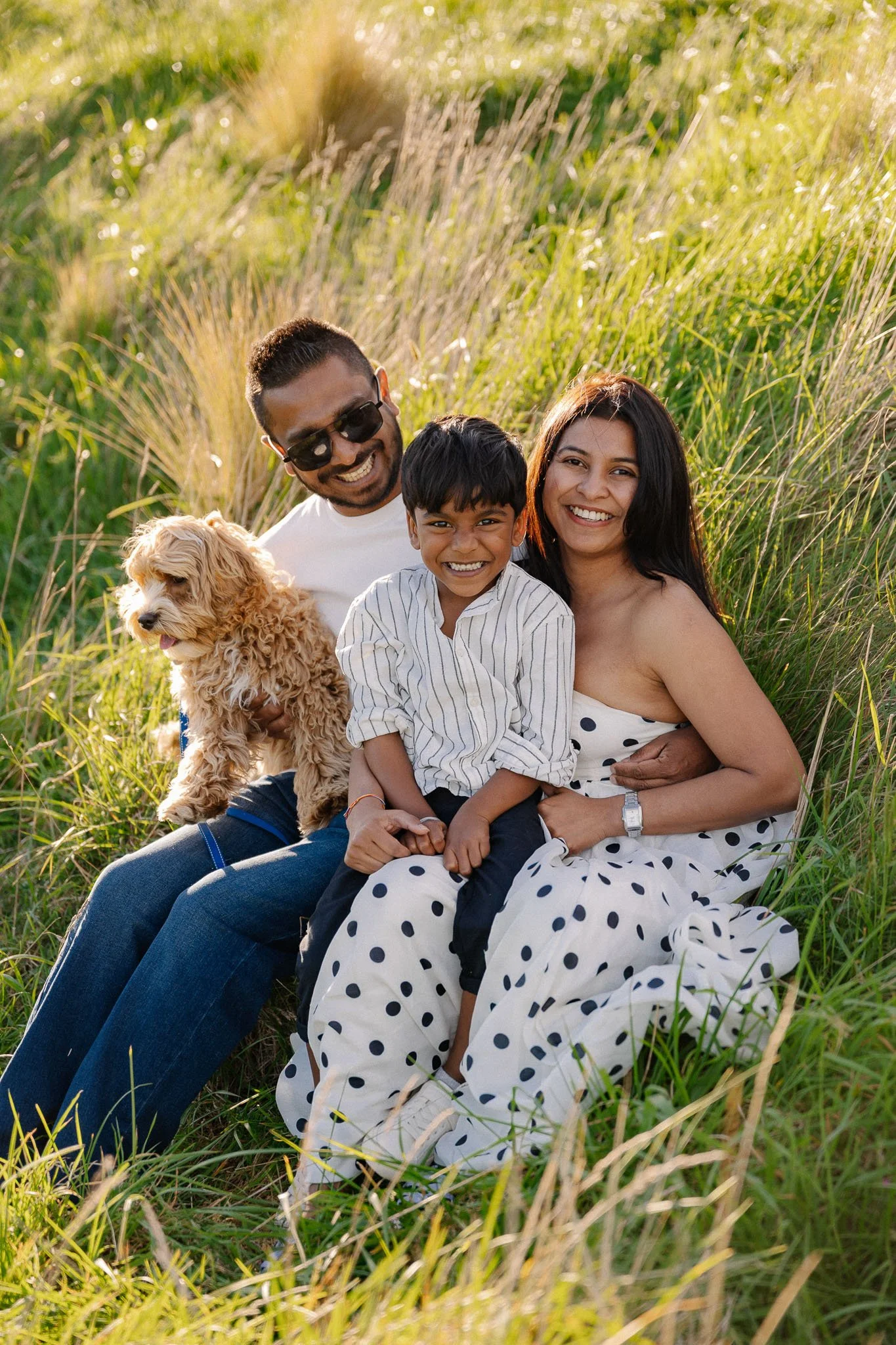 A happy family of three with a dog sitting in tall grass outdoors in sunlight, smiling at the camera.