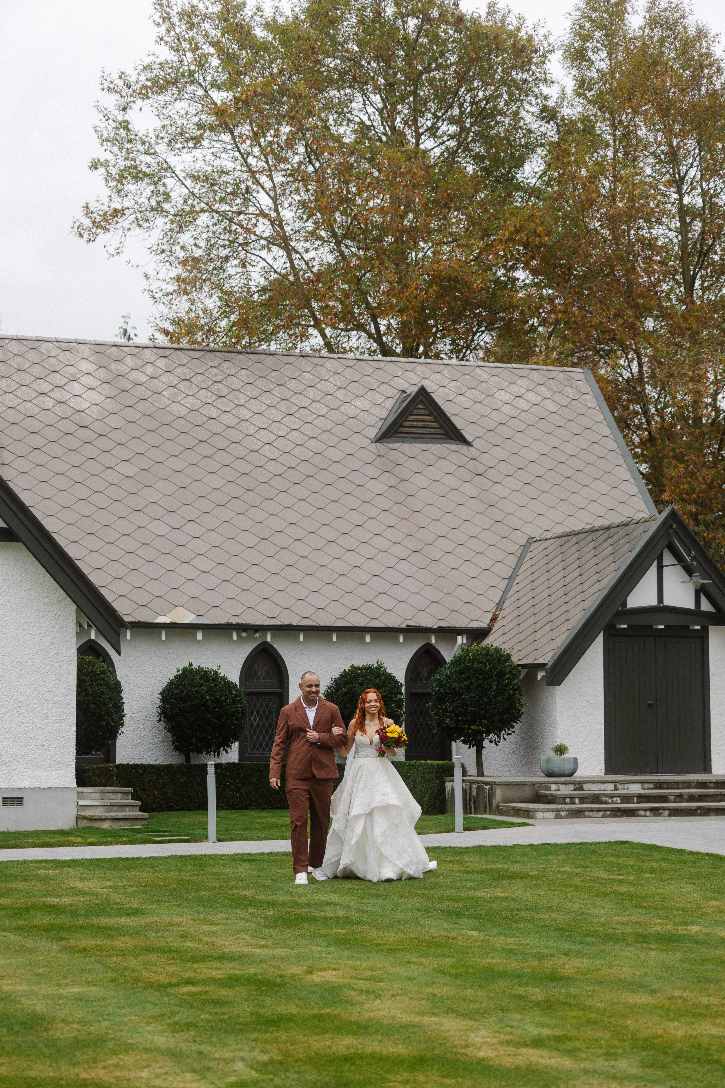 A bride and groom are walking arm in arm outside a white church with black trim, holding a bouquet of colorful flowers. The church has arched windows and a steeply pitched roof, with trees in the background.