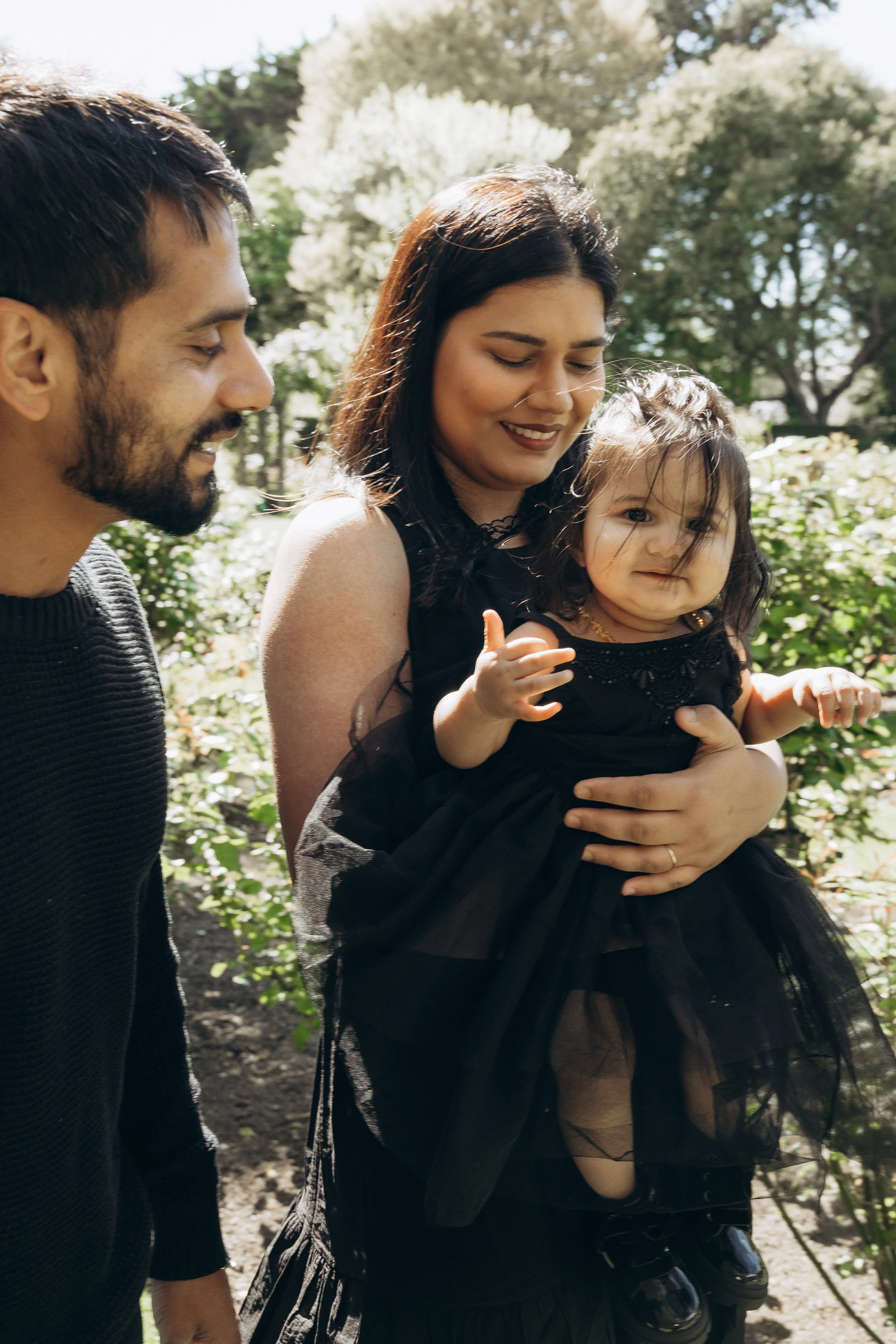 Family with a toddler in a garden, wearing black outfits, surrounded by greenery and trees.