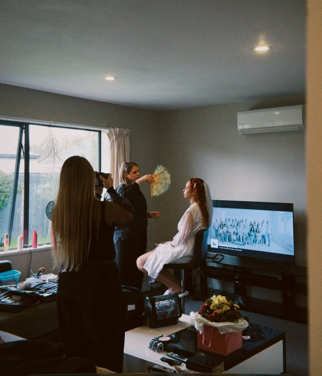 A woman in a wedding dress sitting on a chair with her eyes closed, while a woman holding a fan appears to be doing her makeup or hairstyling. Two other women are standing nearby in a room with a large window, TV, and various items on the table, possibly preparing for a wedding or special event.