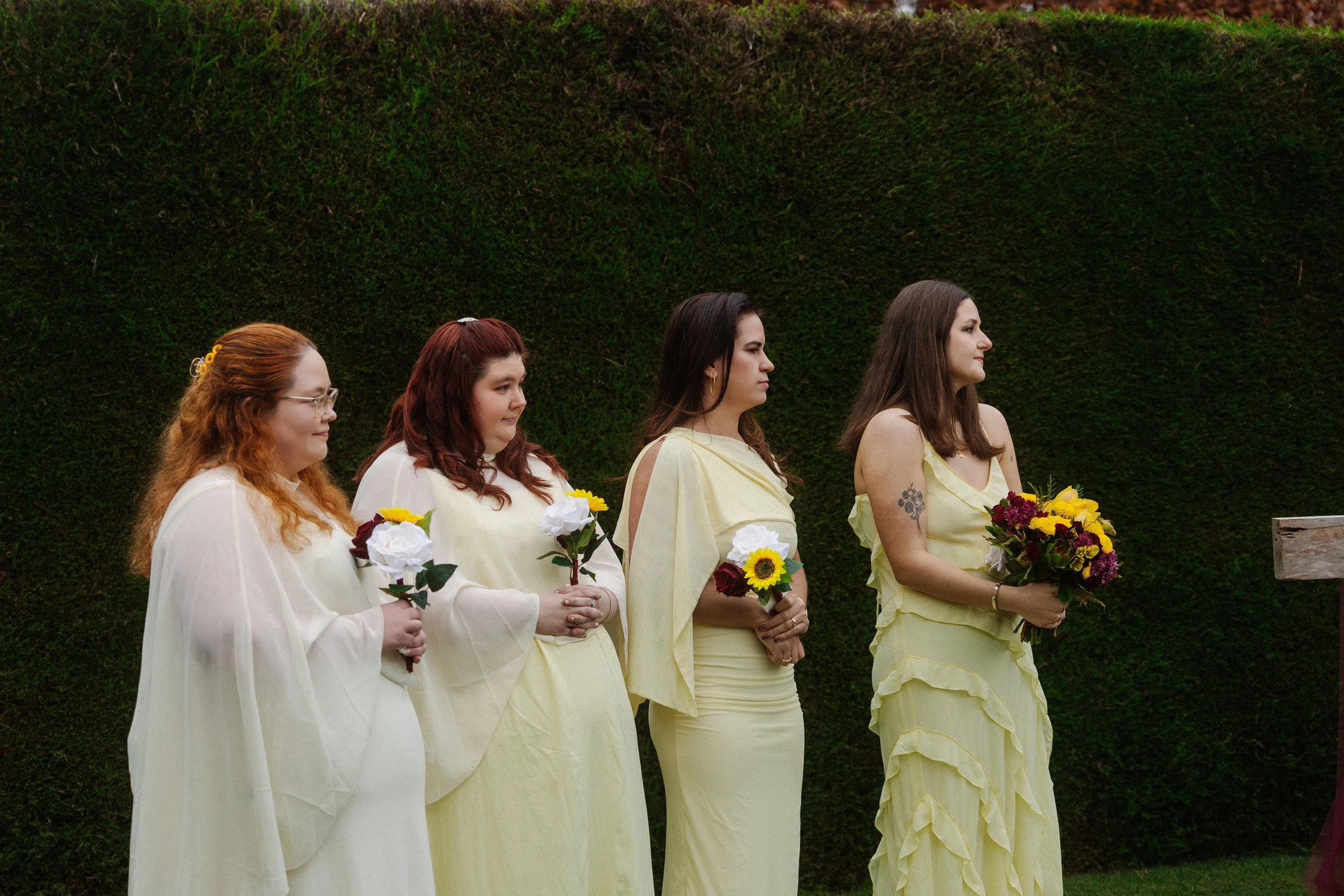 Line of women holding flowers, standing outdoors in front of a green hedge, participating in a ceremony or event.