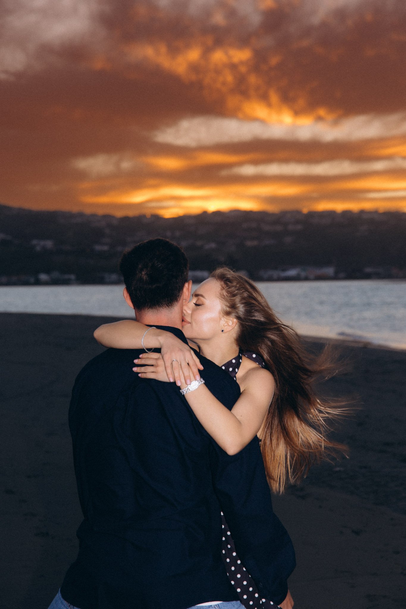 A couple embracing and kissing at sunset on the beach, with the sky filled with orange and dark clouds.
