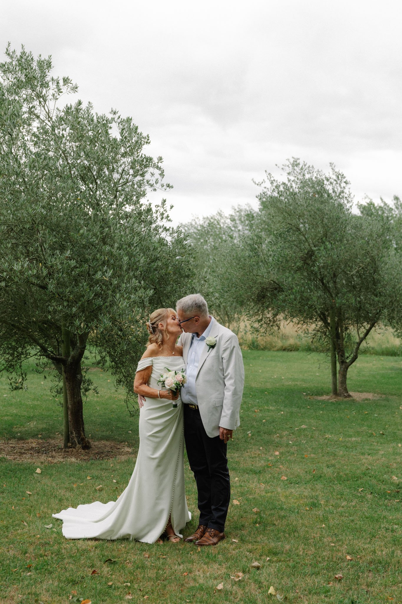 A couple dressed in wedding attire standing outdoors on a grassy area, kissing, with trees and overcast sky in the background.