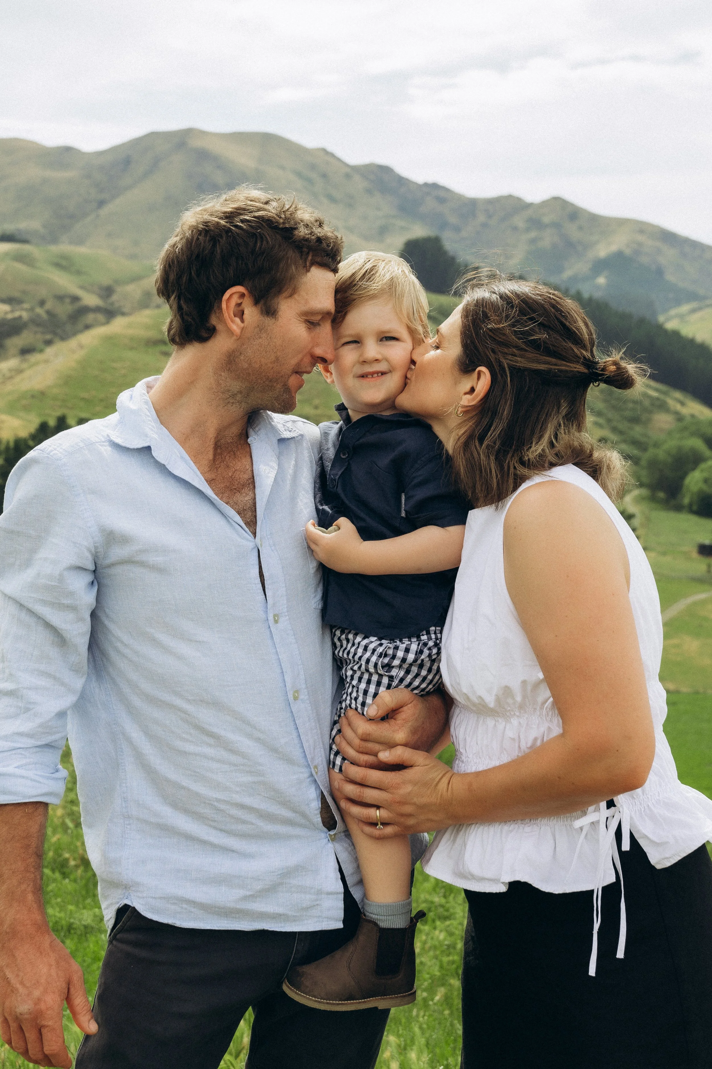 A family of three posing in front of a scenic landscape. A man, woman, and a child. The woman is kissing the child's cheek. The background has rolling green hills and a cloudy sky.