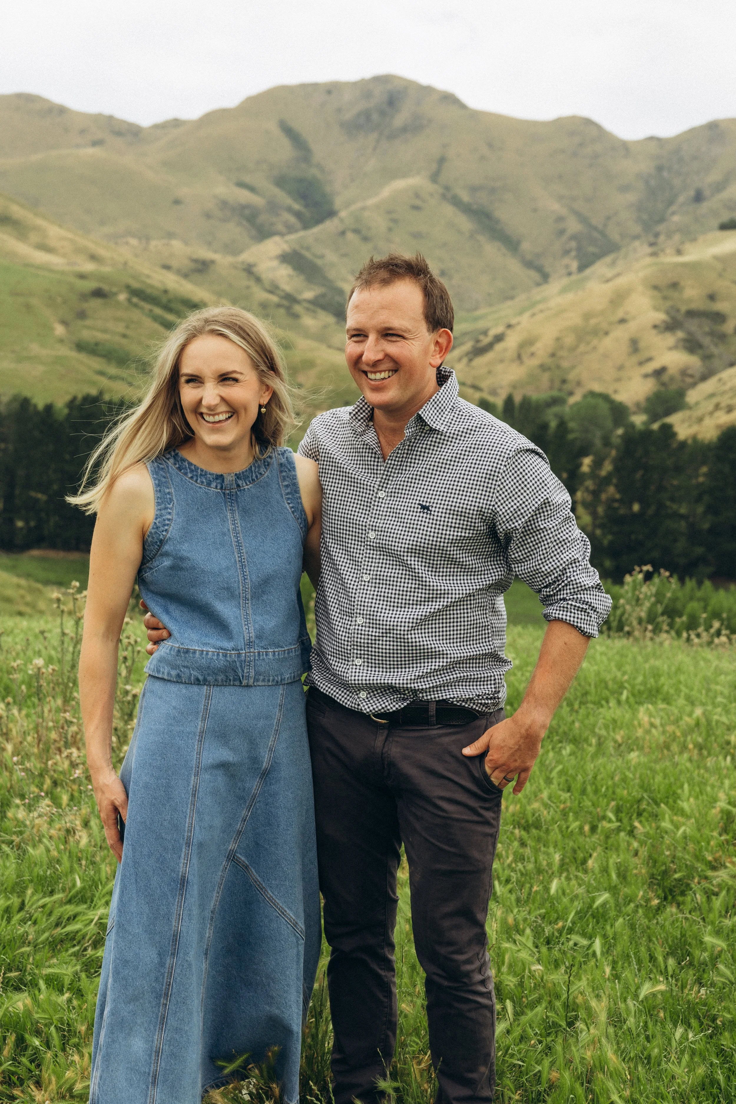 A smiling couple standing in a grassy field with green hills and mountains in the background.