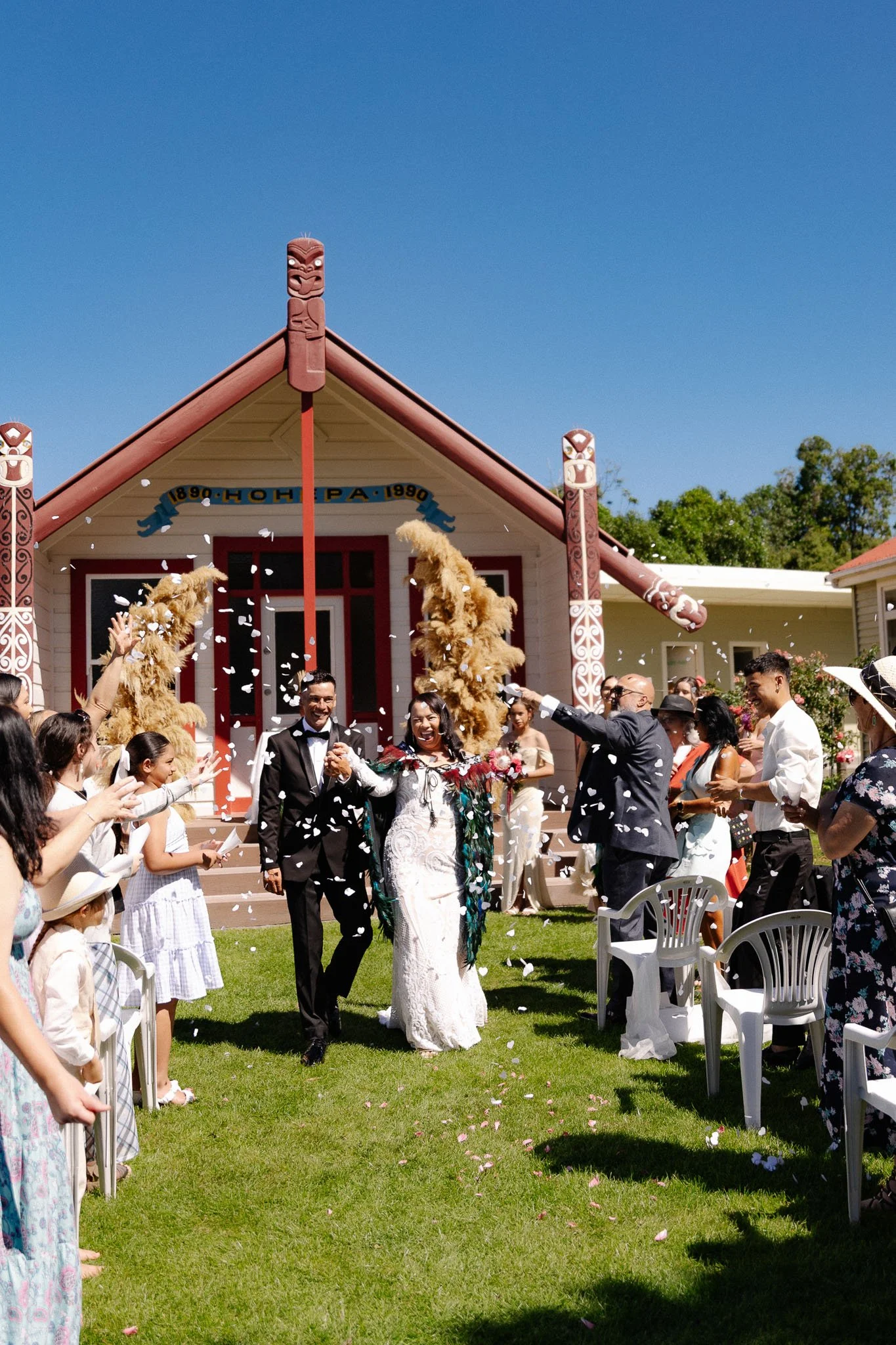 A wedding celebration taking place outdoor in front of a traditional Maori meeting house, with the bride and groom walking down the aisle surrounded by guests, including children, as flower petals are thrown.