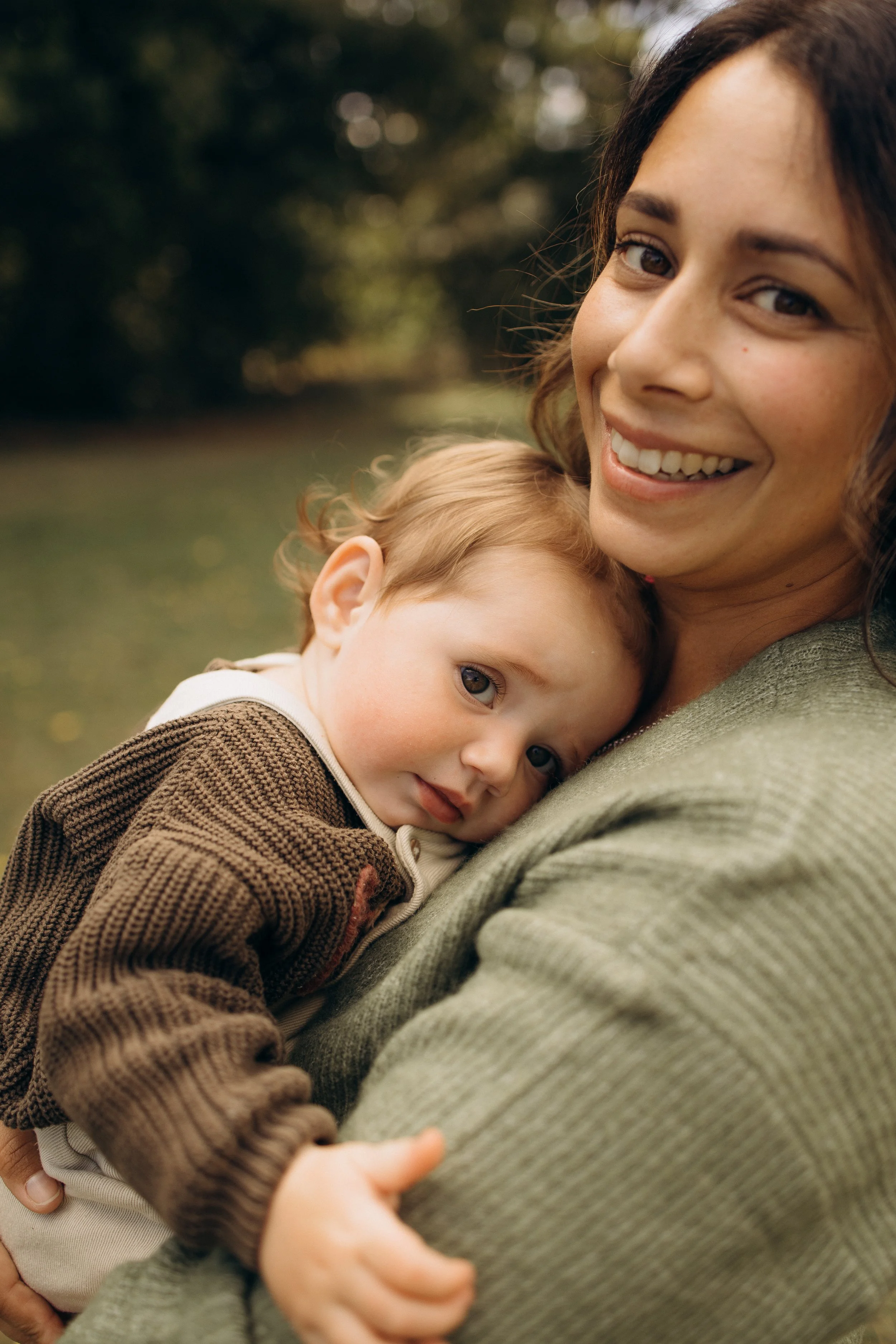 A woman holding a baby close in a park, both smiling, with the baby wearing a brown knitted sweater.
