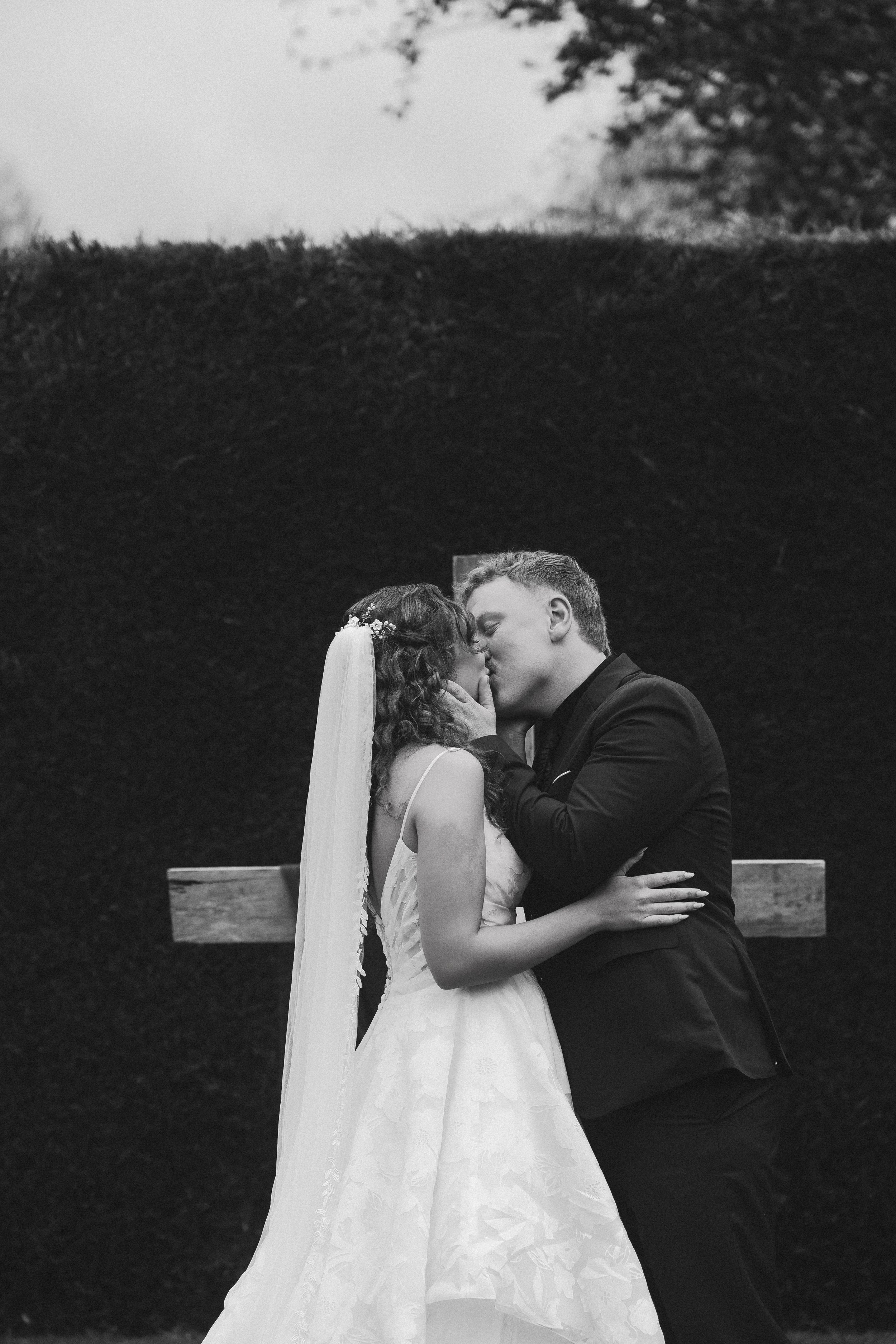 A black and white photo of a bride and groom kissing, standing closely together outdoors. The bride is wearing a wedding gown with lace details and a veil, while the groom is in a suit. They are holding each other tenderly with a hedge and tree in th